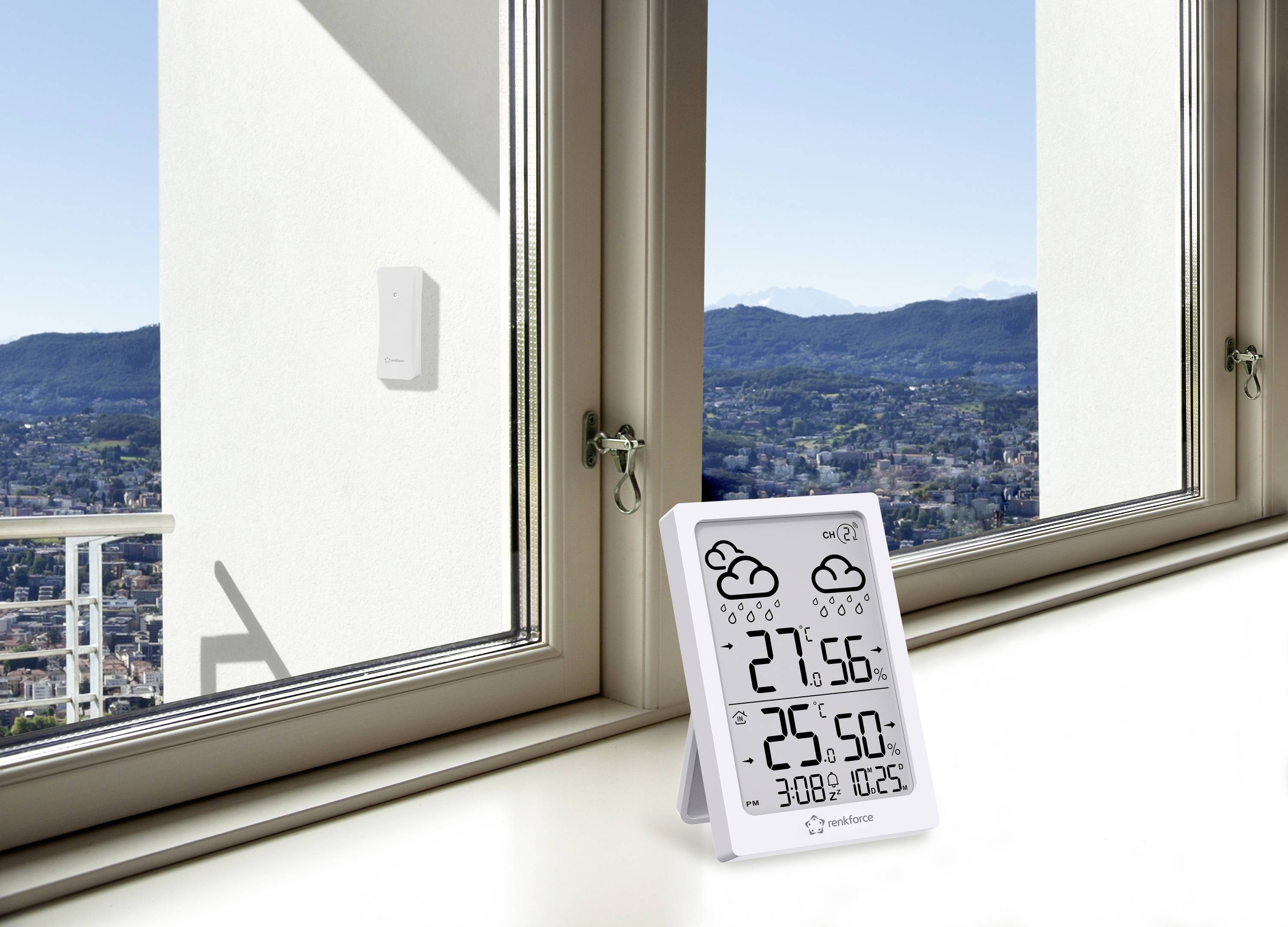 Een thermometer toont op een display binnentemperatuur (27,5°C), buitentemperatuur (25,6°C) en luchtdruk (1012,5 hPa), voor een raam met uitzicht.