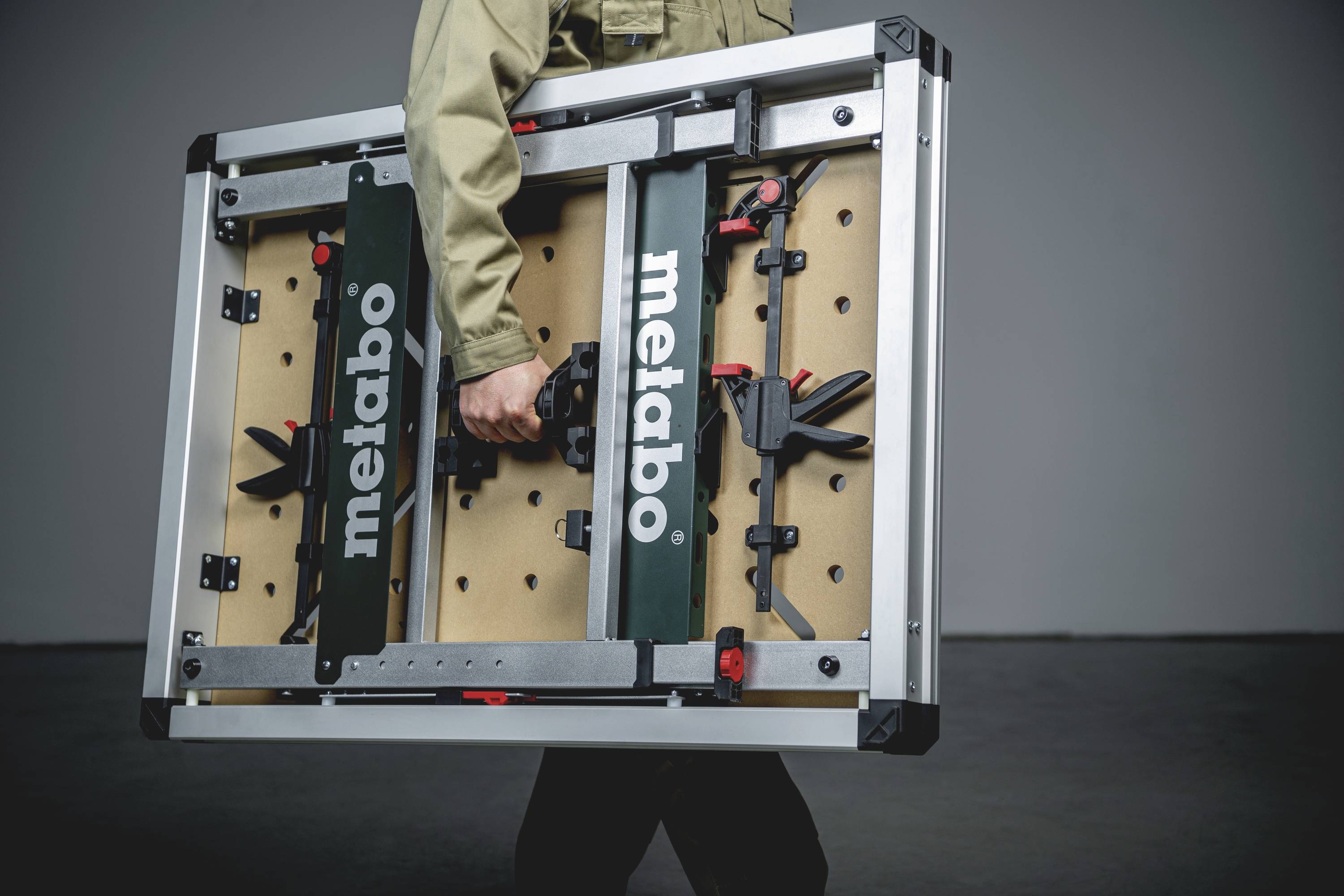 Een persoon draagt een draagbare werktafel van het merk 'Metabo'. De tafel heeft een metalen frame en is opvouwbaar.