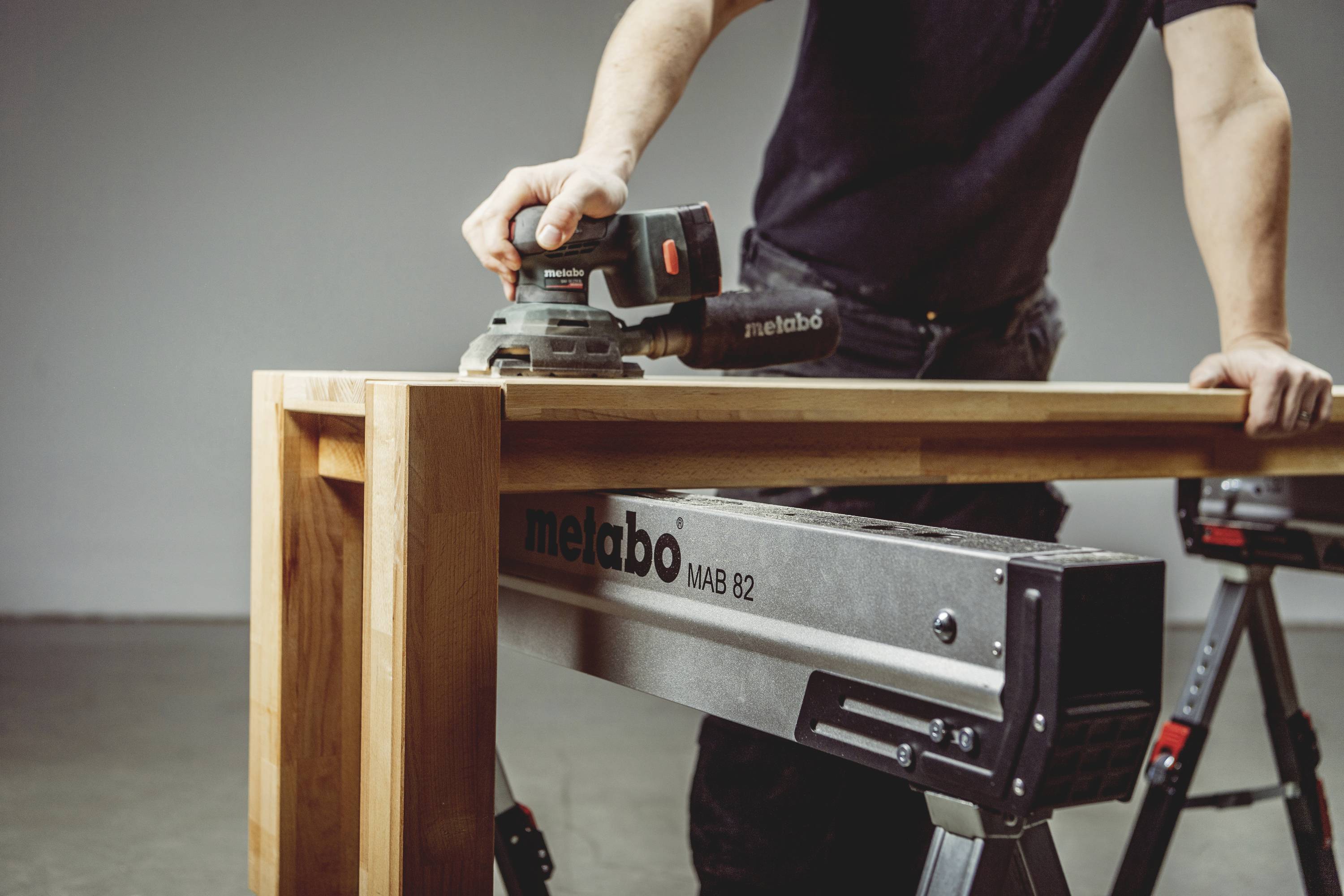 Een persoon schuurt een houten plank met een elektrische schuurmachine op een werkbank. De merknaam van het gereedschap is zichtbaar.