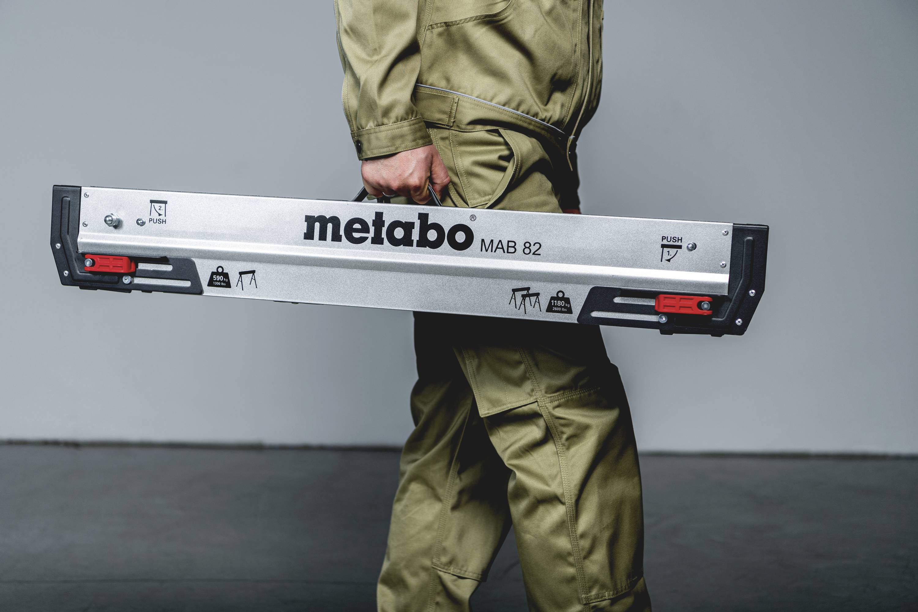 Een persoon draagt een zilveren gereedschapskist met het opschrift 'metabo MAB 82'. De achtergrond is neutraal.