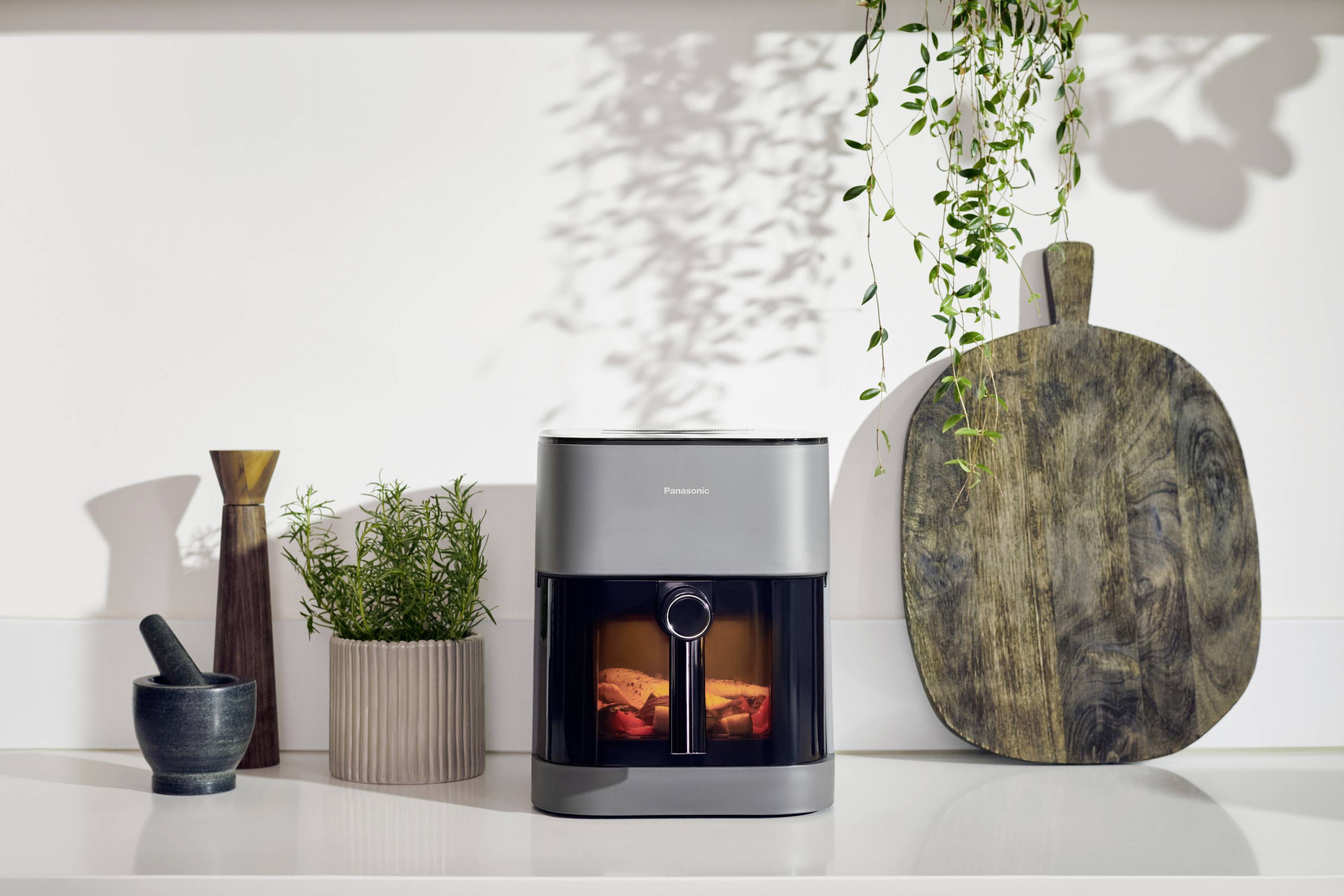 Moderne keuken met grijze heteluchtfriteuse, omringd door kruiden, een vijzel en een snijplank; decoratieve plant hangt rechtsboven.