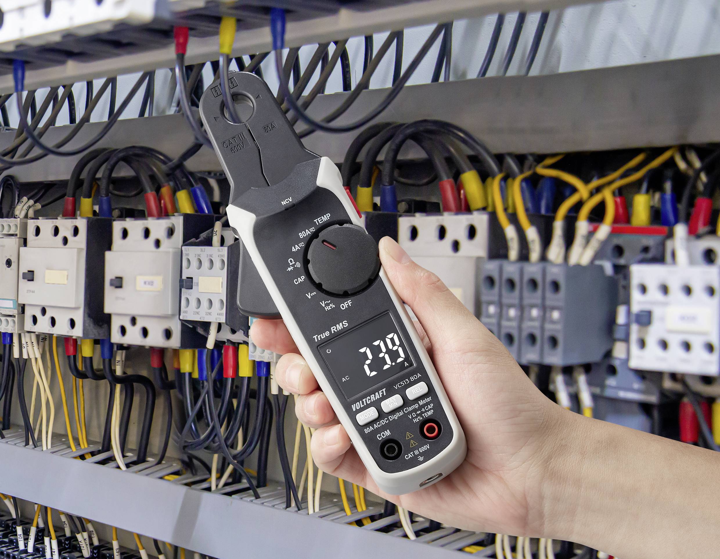 Een hand houdt een digitale multimeter voor een elektrisch paneel met draden en schakelaars, waarbij een aflezing van 23,9 wordt weergegeven.