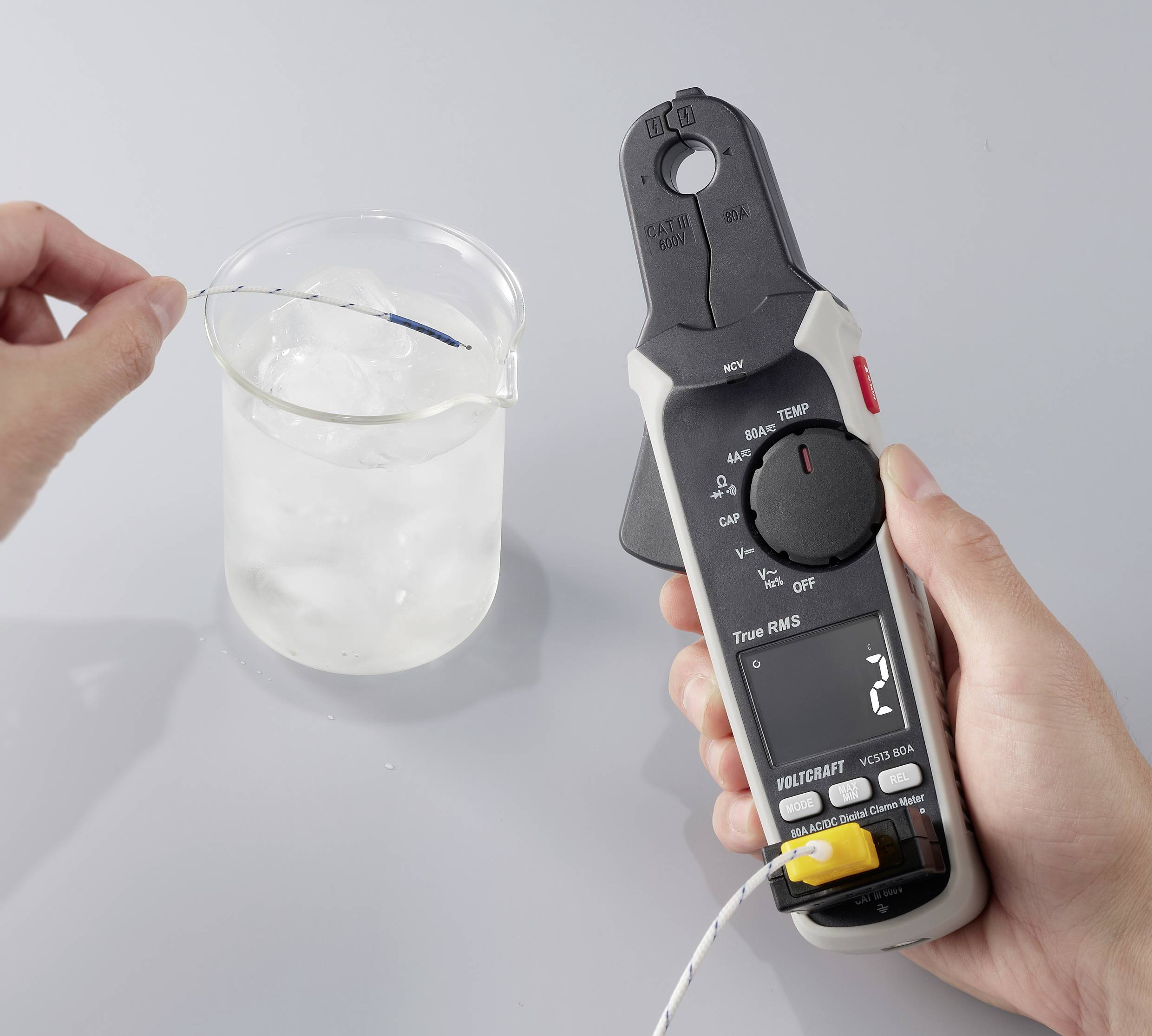 Een persoon gebruikt een digitale thermometer om de temperatuur van vloeistof in een bekerglas te meten, dat gevuld is met ijswater op een grijze ondergrond.