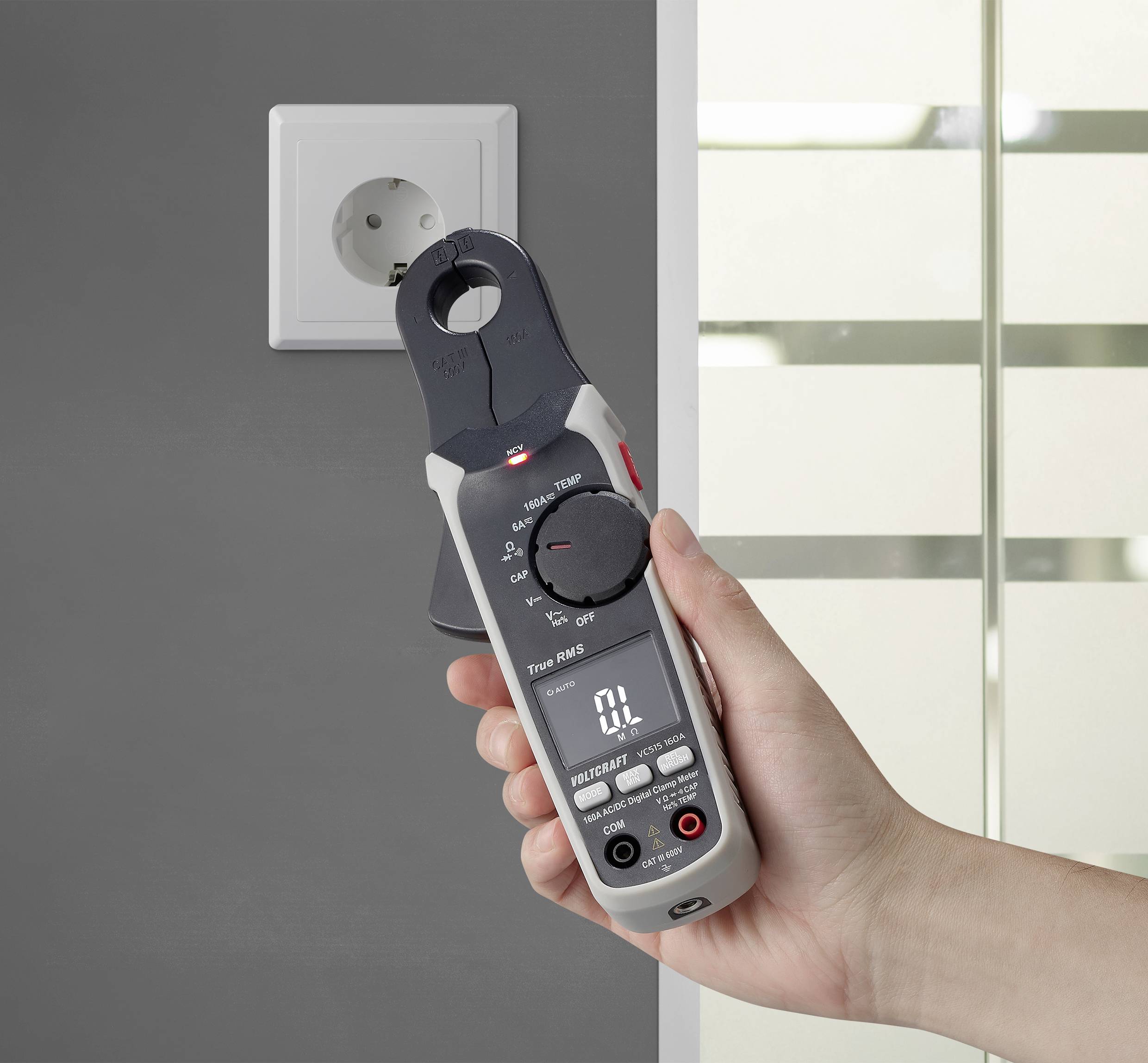 Een hand houdt een digitale multimeter naast een stopcontact aan een grijze muur. Het apparaat toont '0 L' op het display.