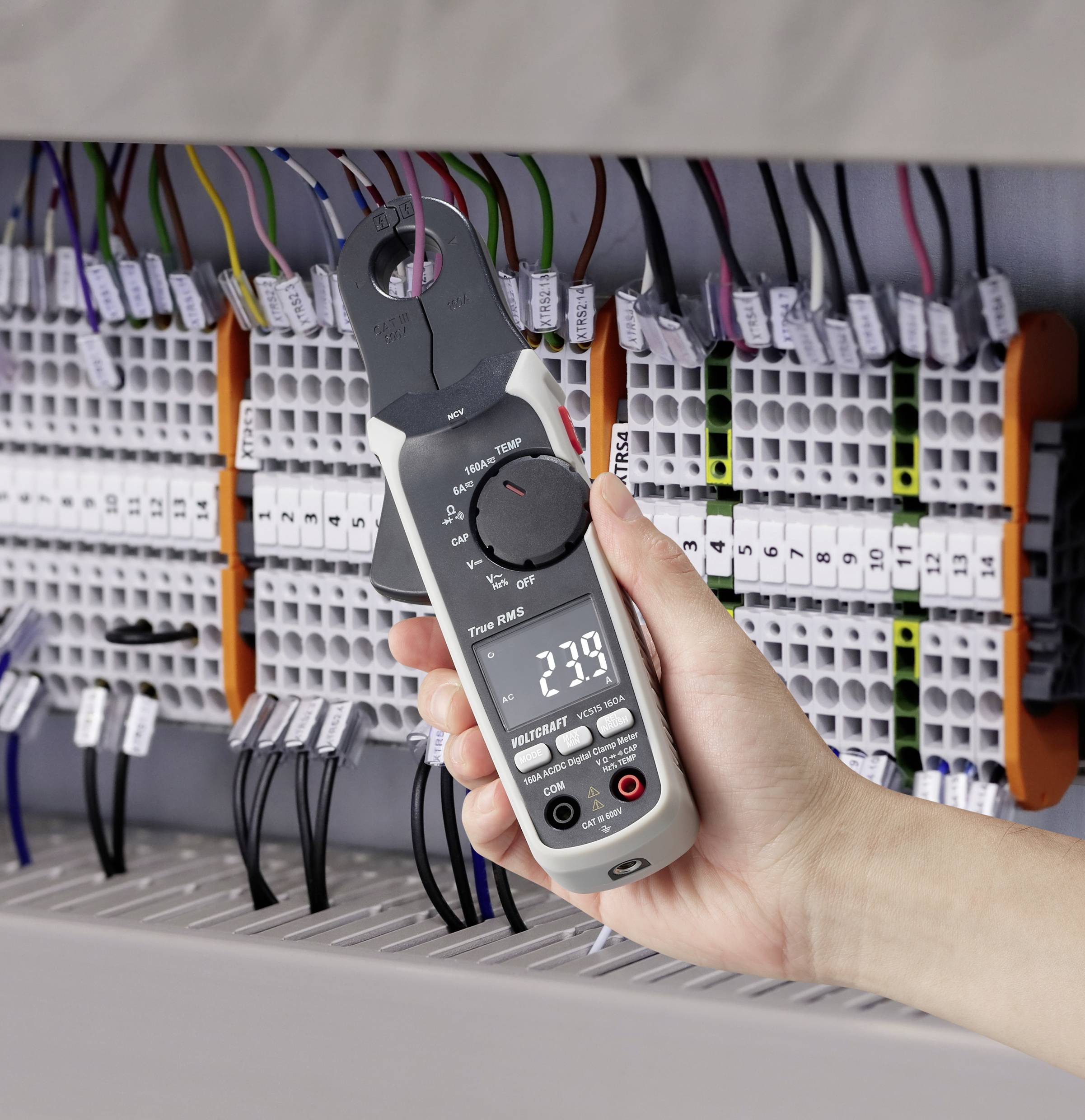 Een hand houdt een digitale stroomtangmeter voor een schakelkast met talrijke kabels. Het scherm toont '239'.