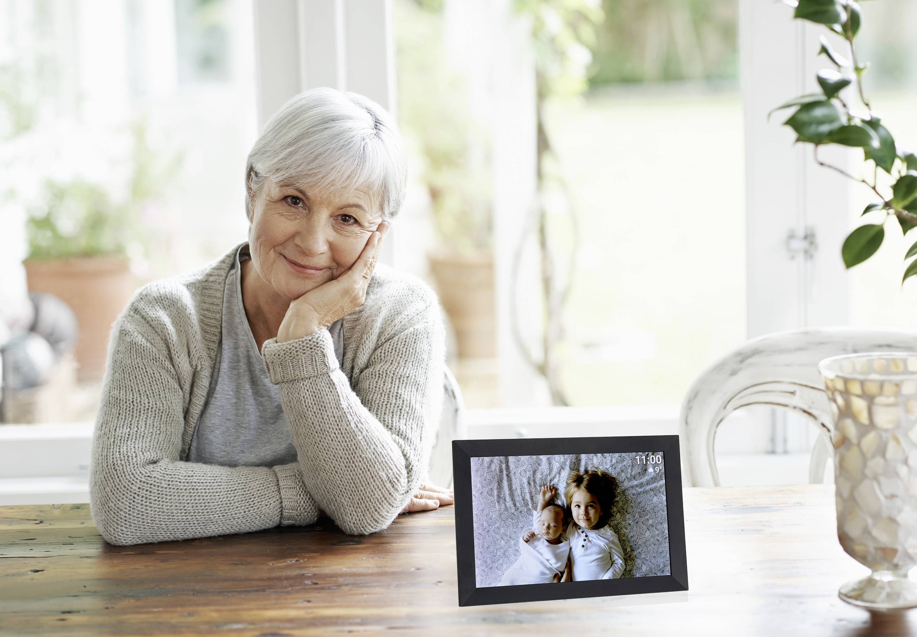 Oudere vrouw glimlacht aan tafel, steunt haar gezicht met de hand. Op tafel staat een digitale fotolijst met twee kinderen.