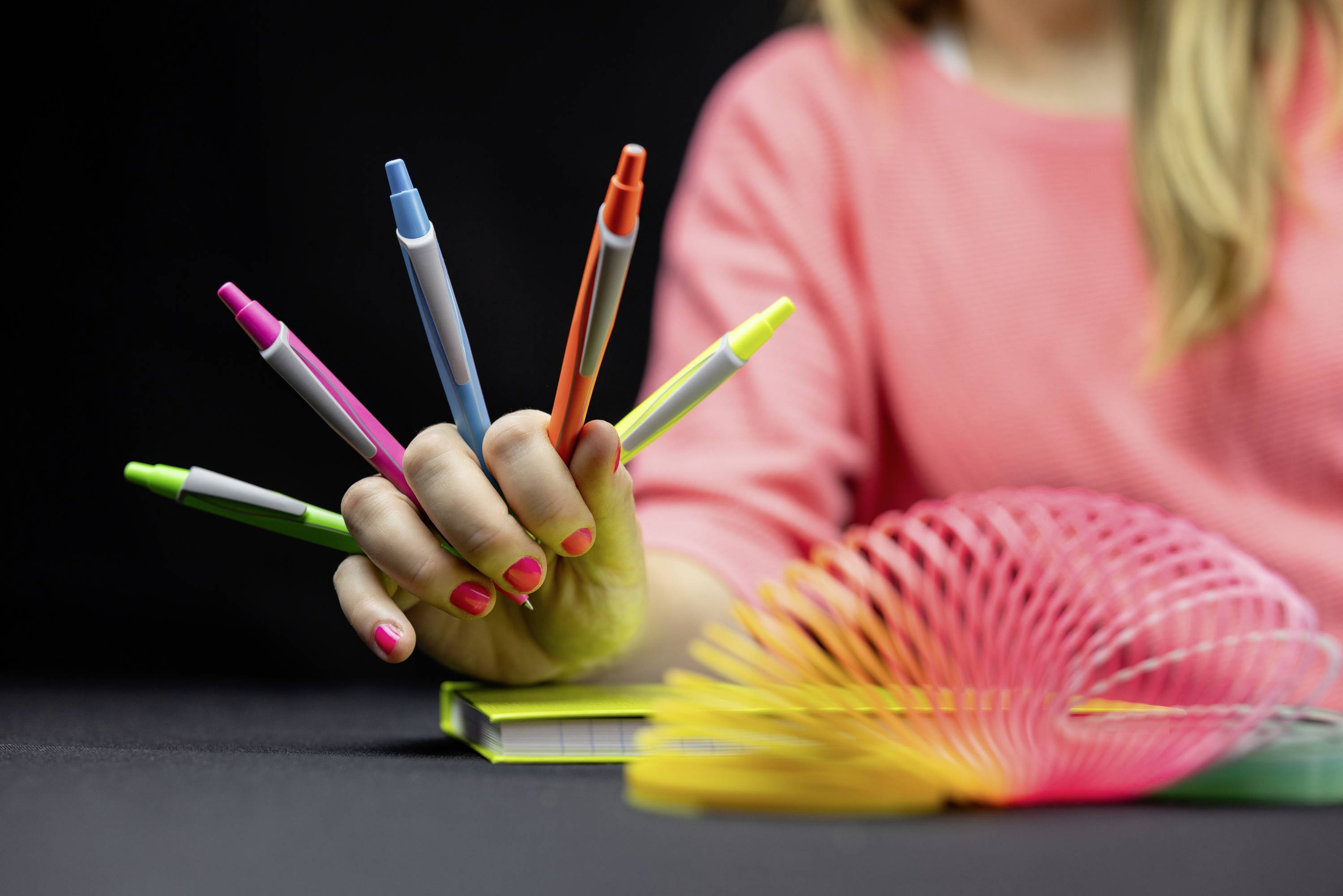 Een persoon houdt vijf kleurrijke pennen tussen de vingers op een speelse manier. Op de voorgrond is een kleurrijke spiraal-slinky te zien.