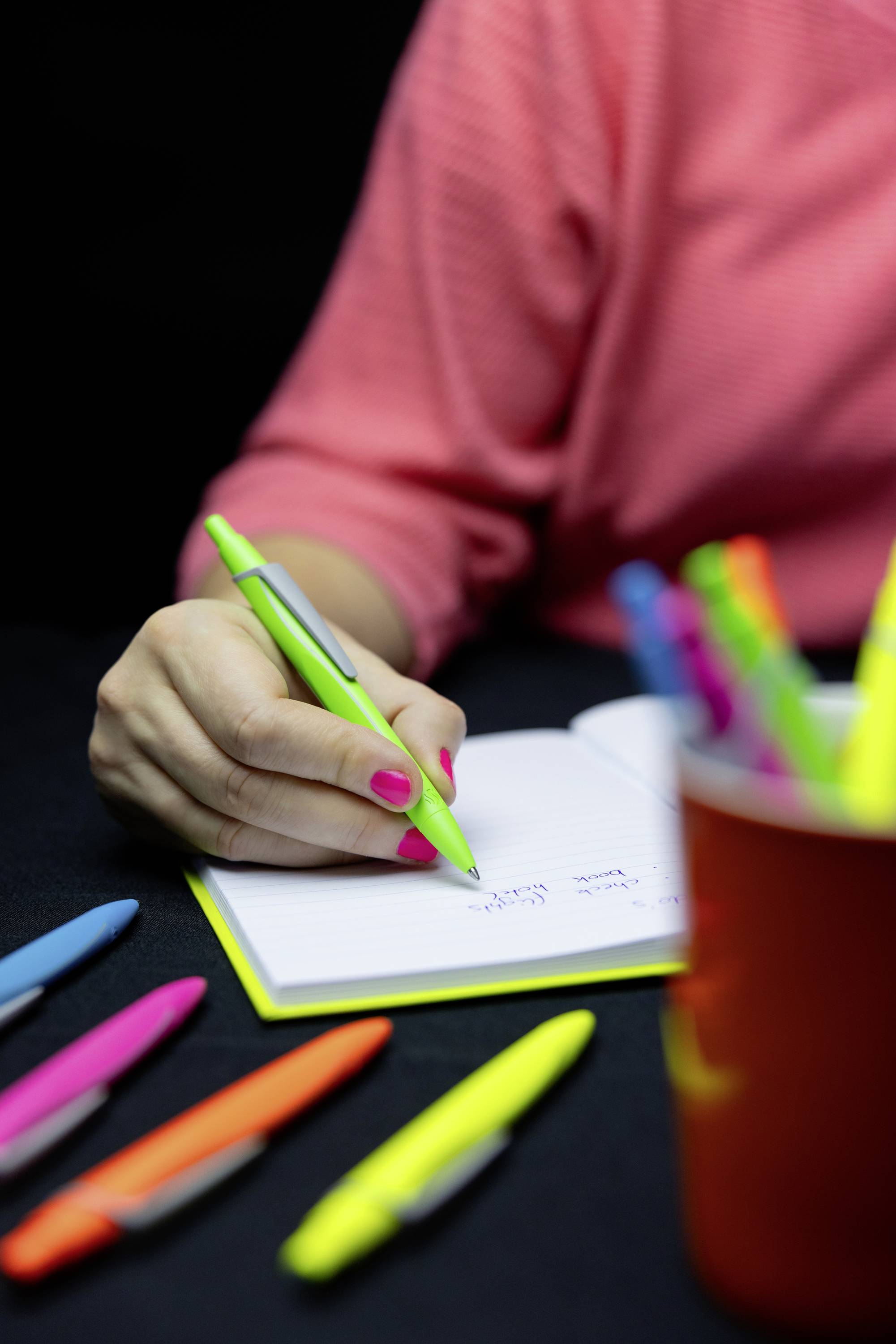 Een persoon schrijft met een groene pen in een notitieboek. Op de voorgrond liggen verschillende gekleurde pennen verspreid, en een beker met nog meer pennen is gedeeltelijk zichtbaar.