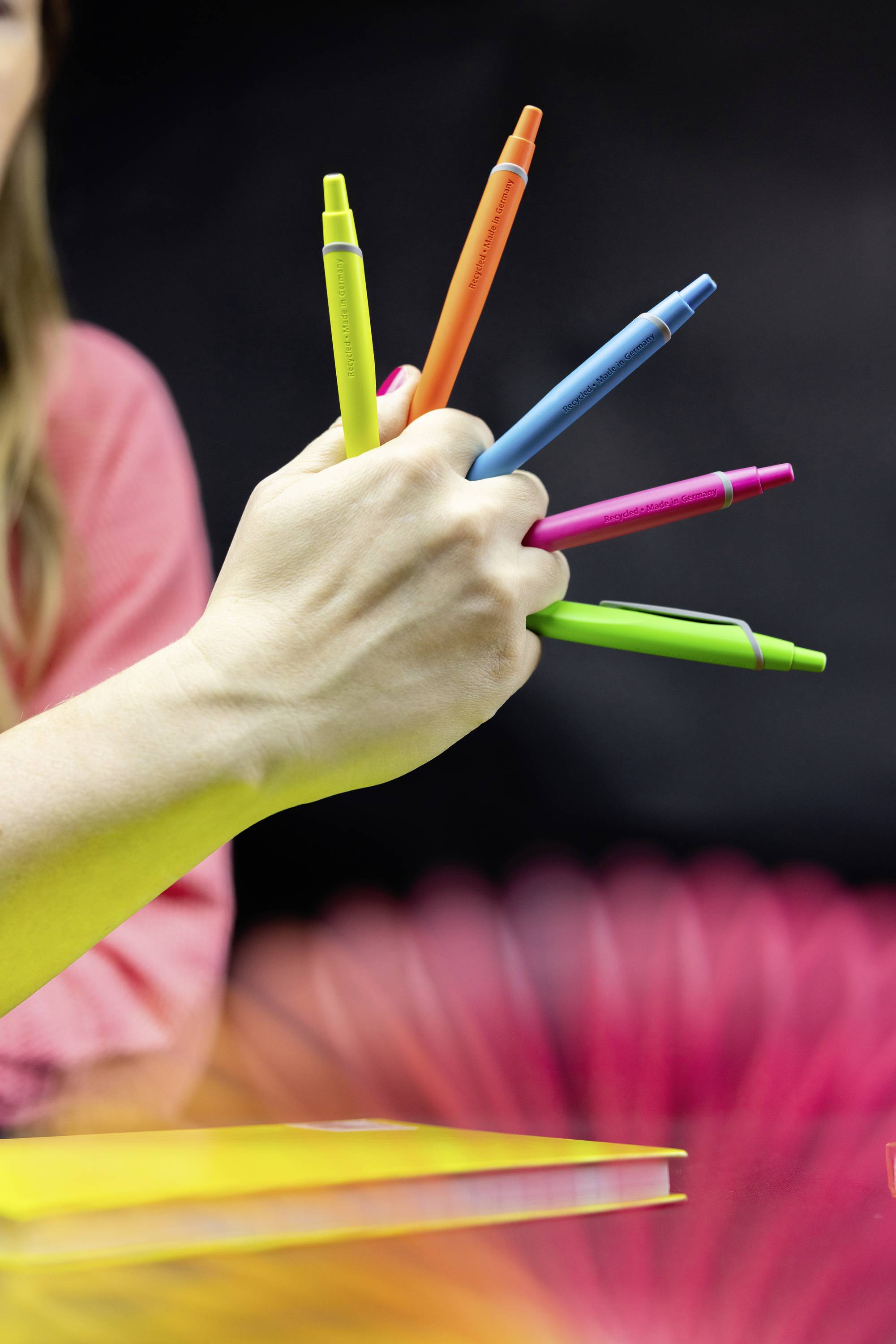Een hand houdt vijf kleurrijke pennen vast, uitwaaierd als een waaier; onscherpe achtergrond met een rood en zwart kleurverloop.