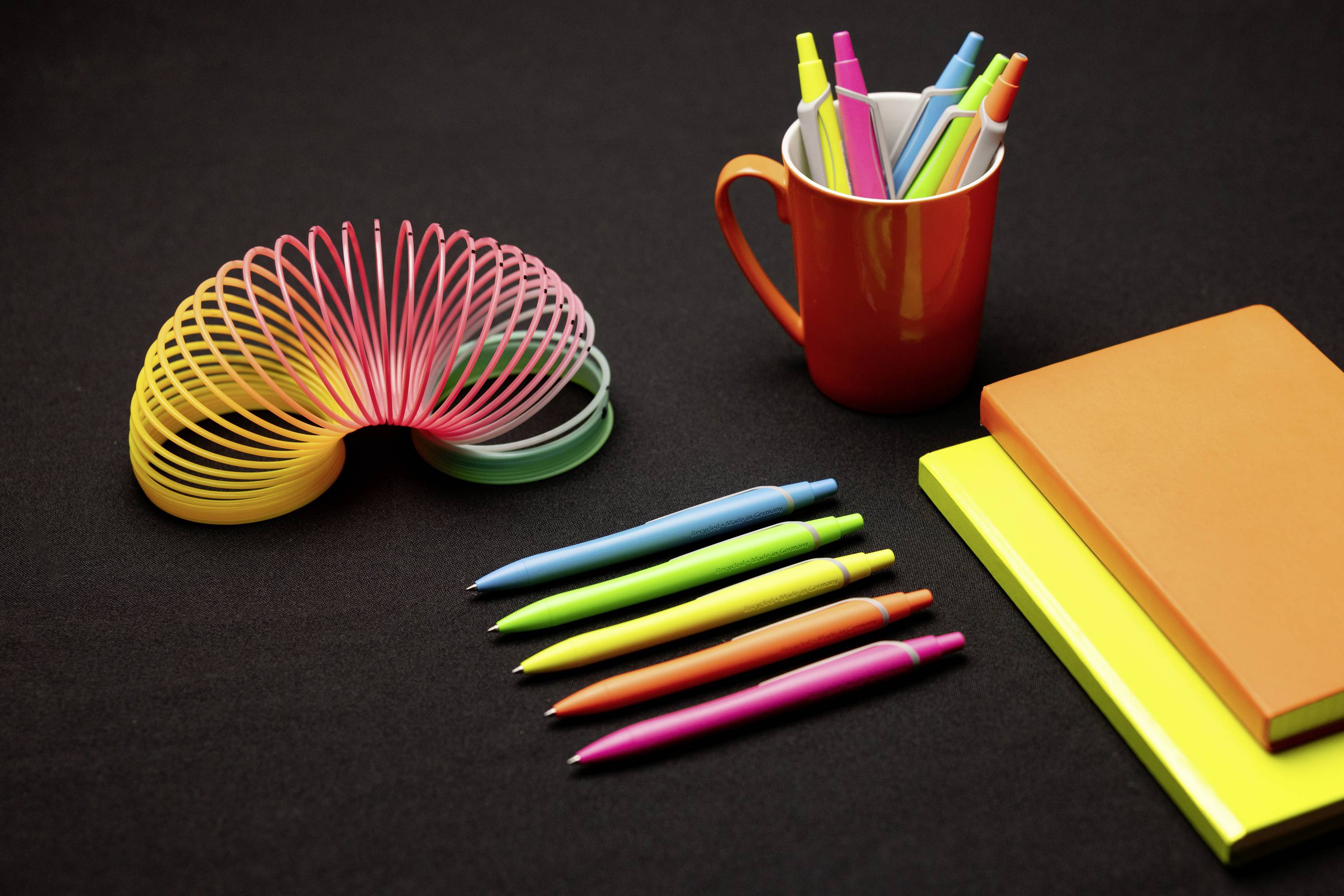 Kleurrijke kantoorartikelen op een zwarte achtergrond: pennen, een rode mok met markers, een regenboog-slinky, twee notitieblokken.