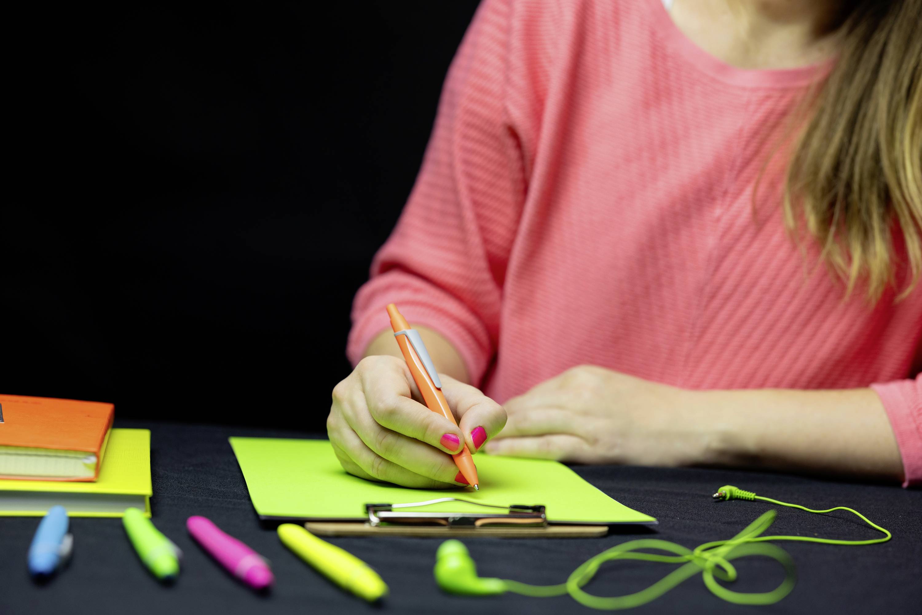 Een persoon in een roze bovenkleding schrijft met een oranje pen op lichtgroen papier. Verschillende pennen liggen op tafel.