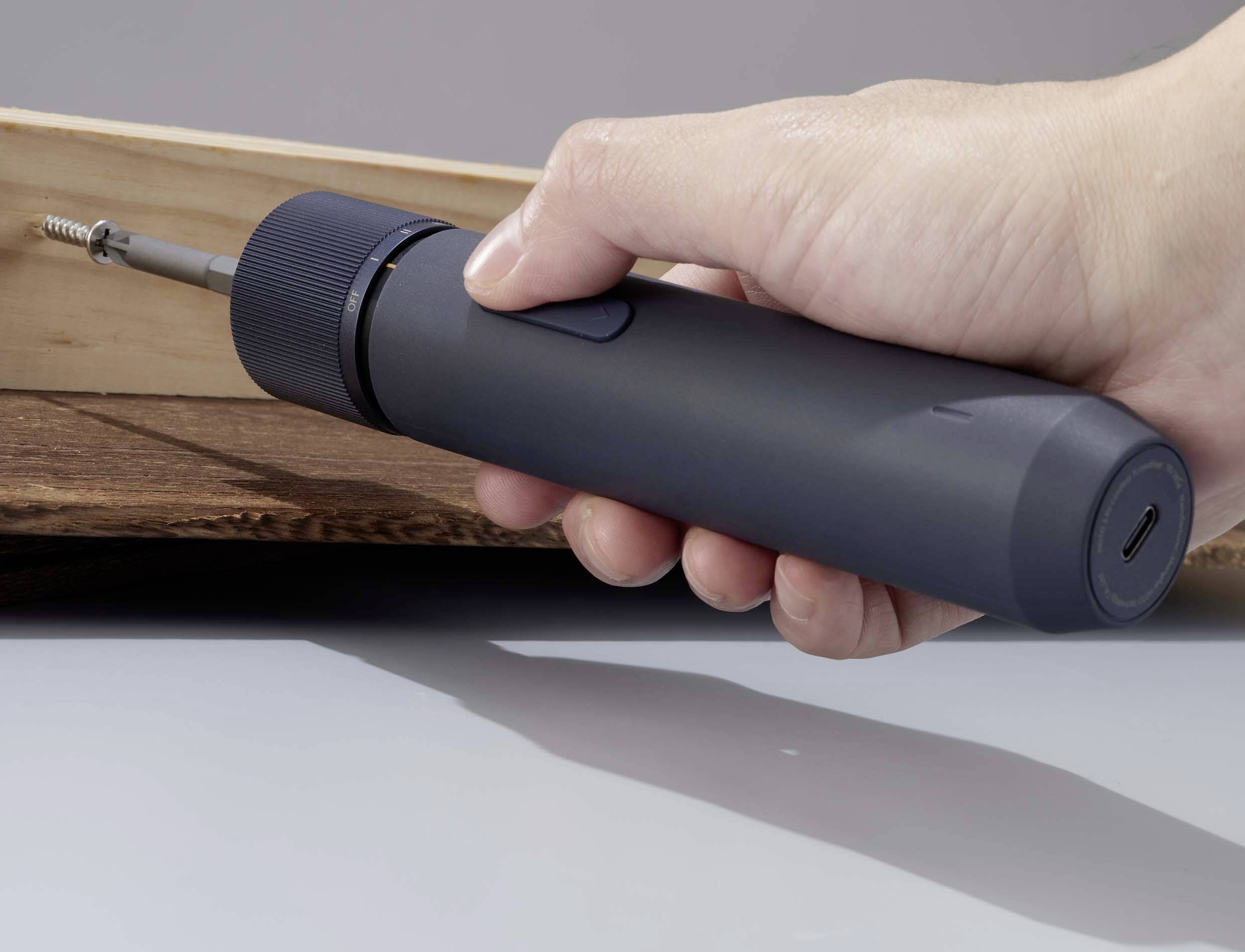 Een hand houdt een elektrische schroevendraaier vast die een schroef in een houten plank draait. De schroevendraaier is donkergrijs.