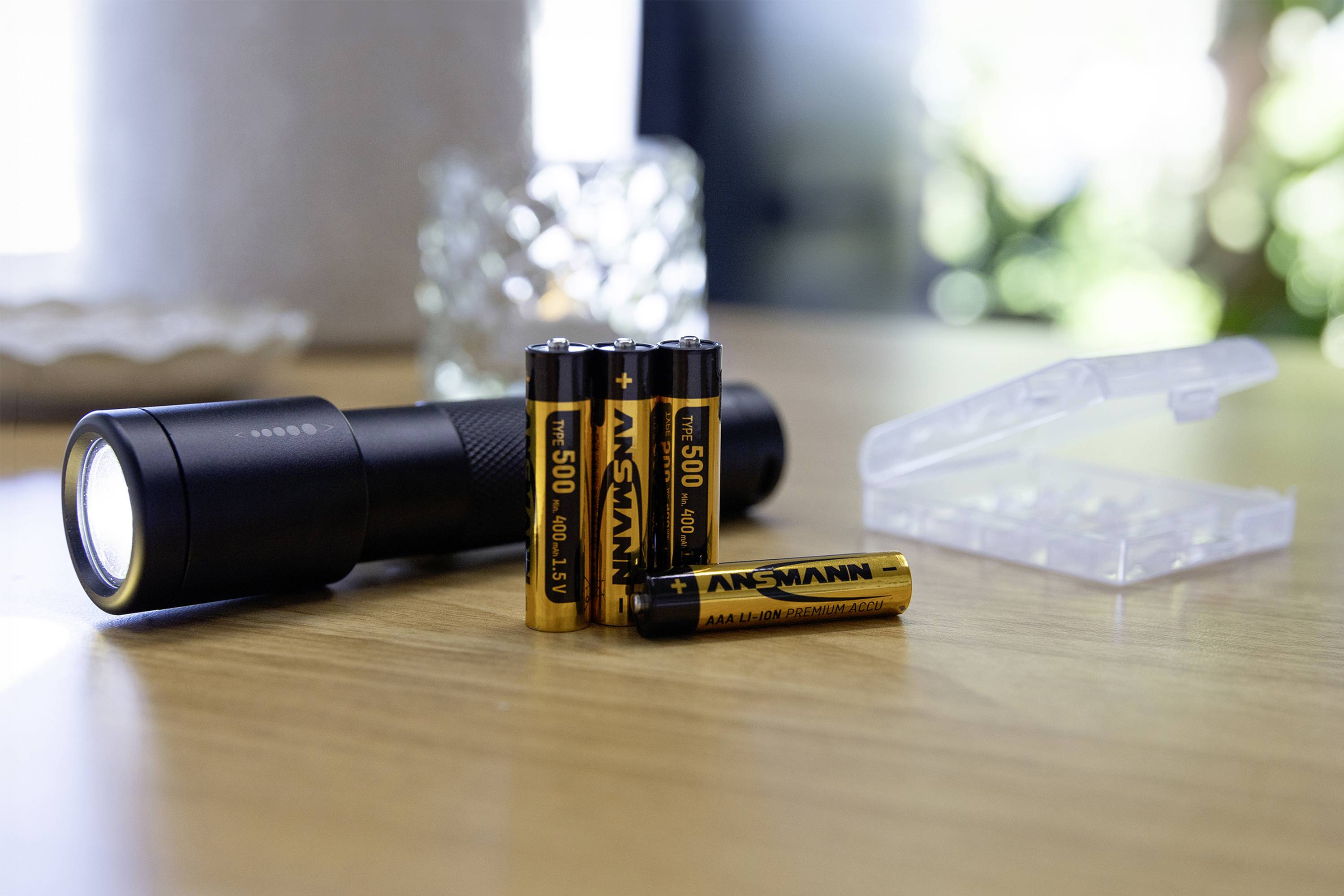 Zaklamp met meerdere batterijen op een houten tafel, batterijen zijn van het merk Ansmann, batterijvak open op de achtergrond.