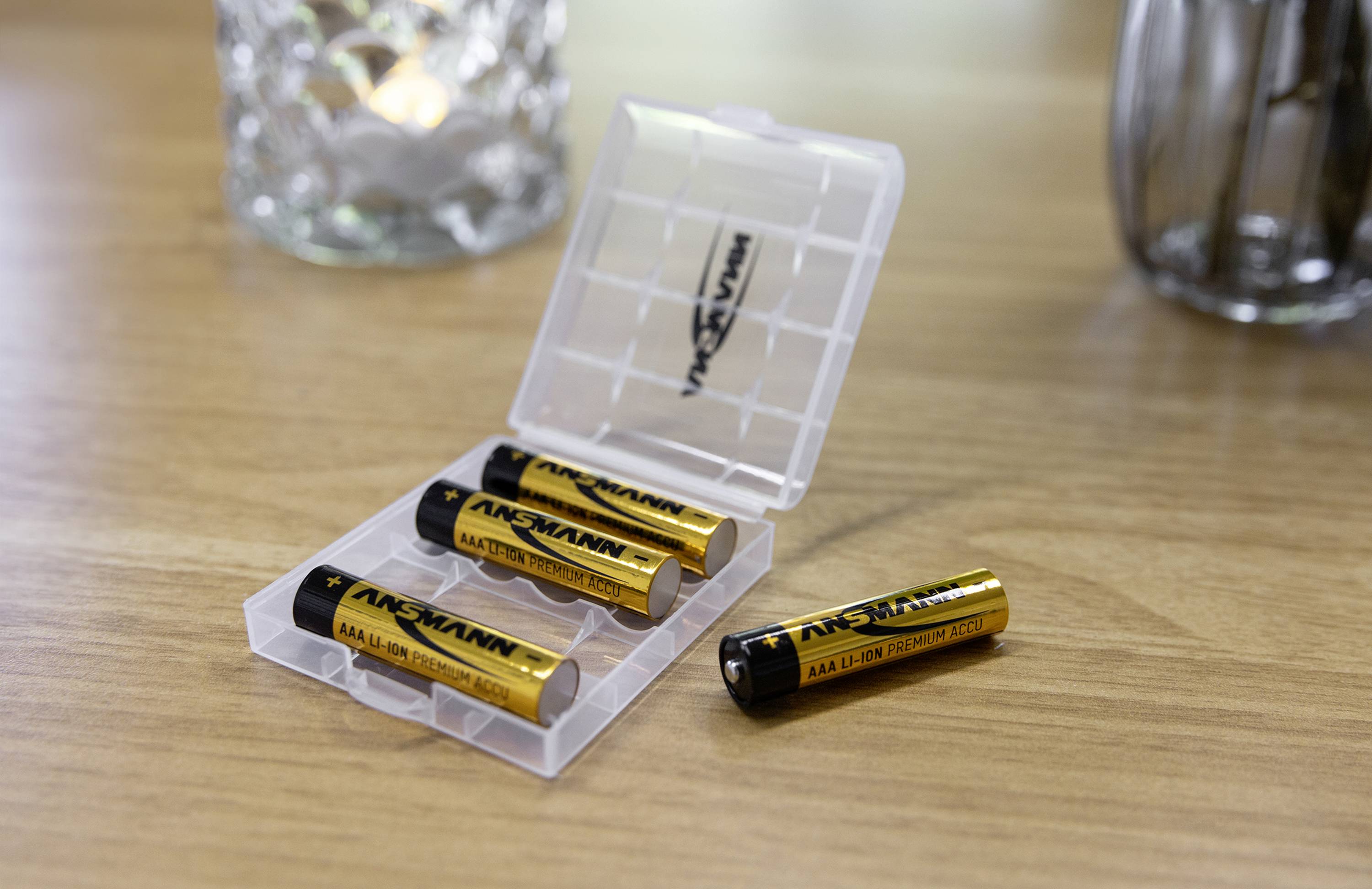 Vier AA-batterijen in een transparante doos op een houten tafel; één batterij ligt ernaast.