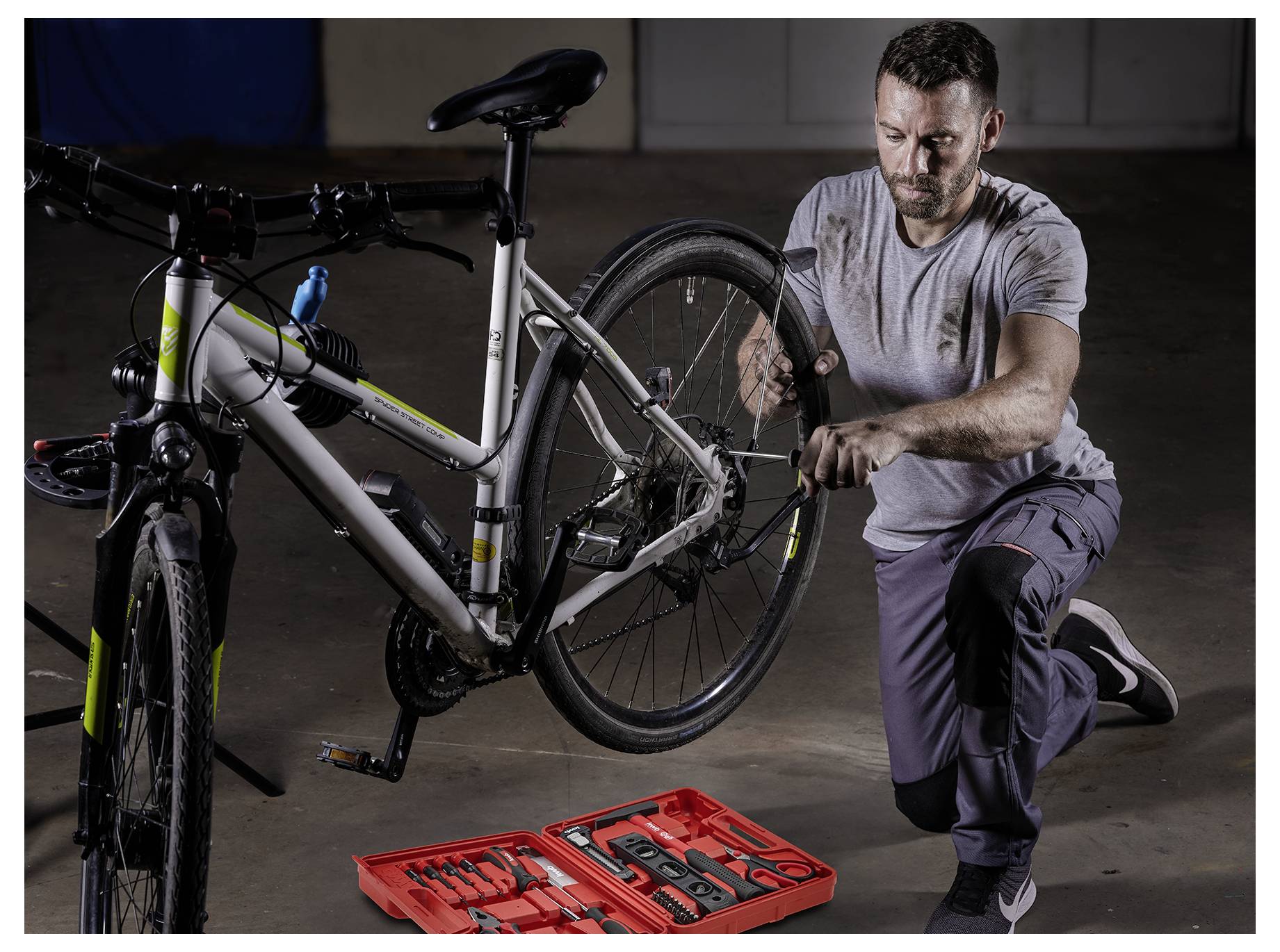 Een man repareert een fietswiel binnenshuis, geknield naast een fiets op een standaard. Een rode gereedschapskoffer staat open op de vloer naast hem.