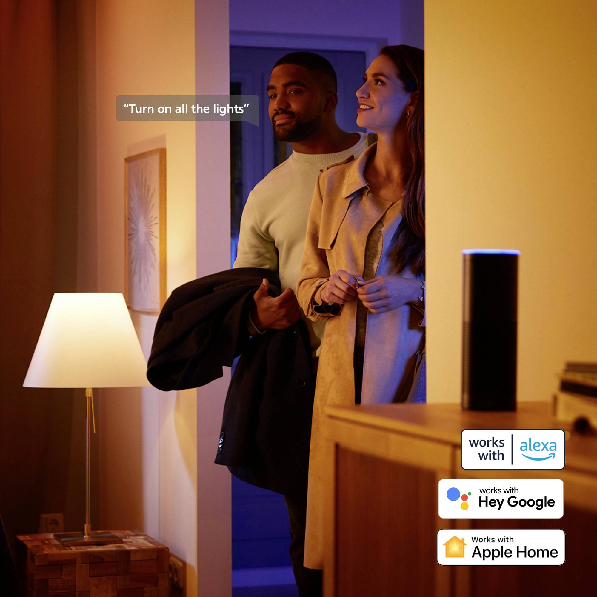 Een stel betreedt lachend een ruimte. Een staande lamp verlicht de scène. Een smart speaker is zichtbaar.