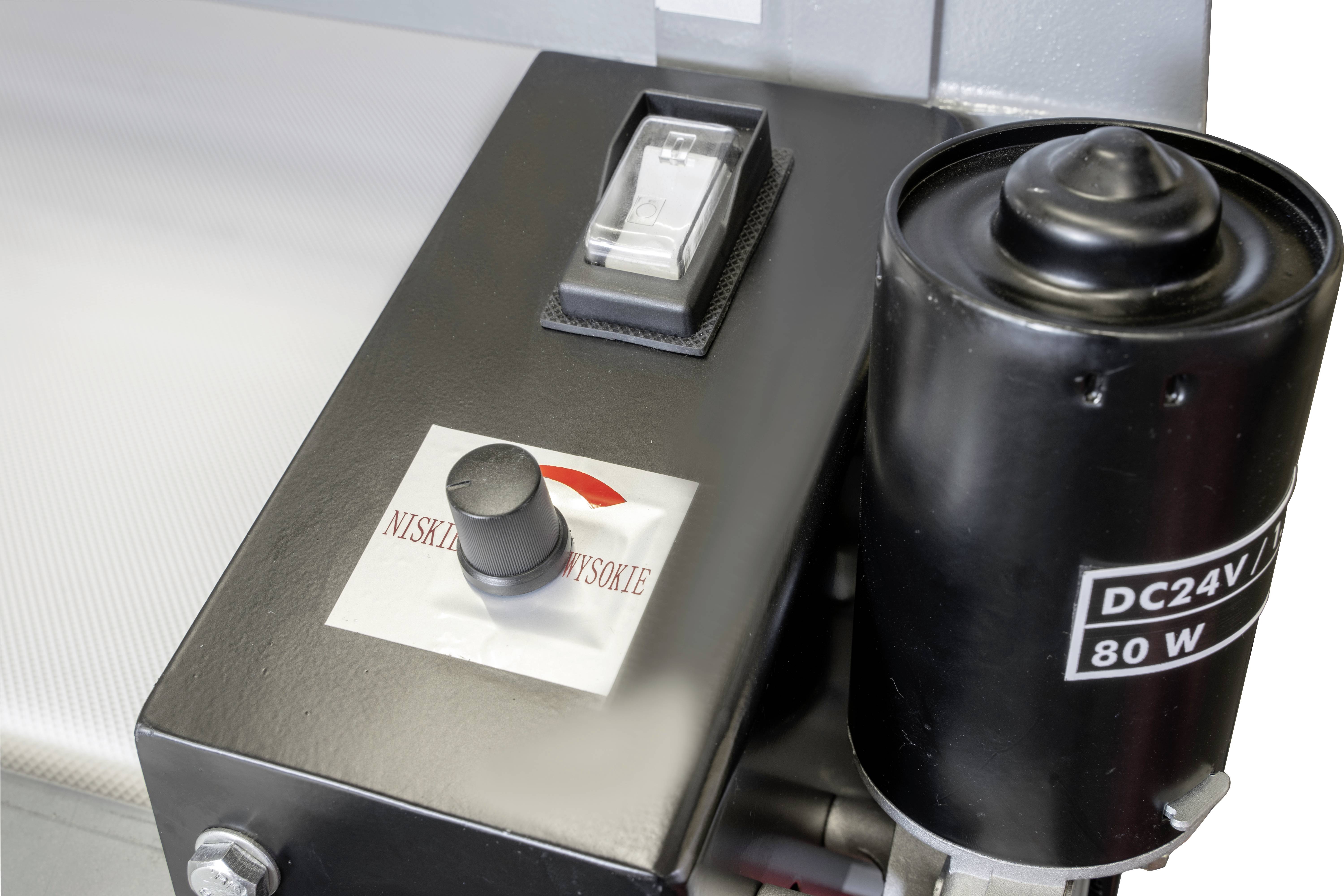 Een zwarte metalen cilinder met het opschrift 'DC24V 80W' en een besturingskast met schakelaar en knop op een machine.