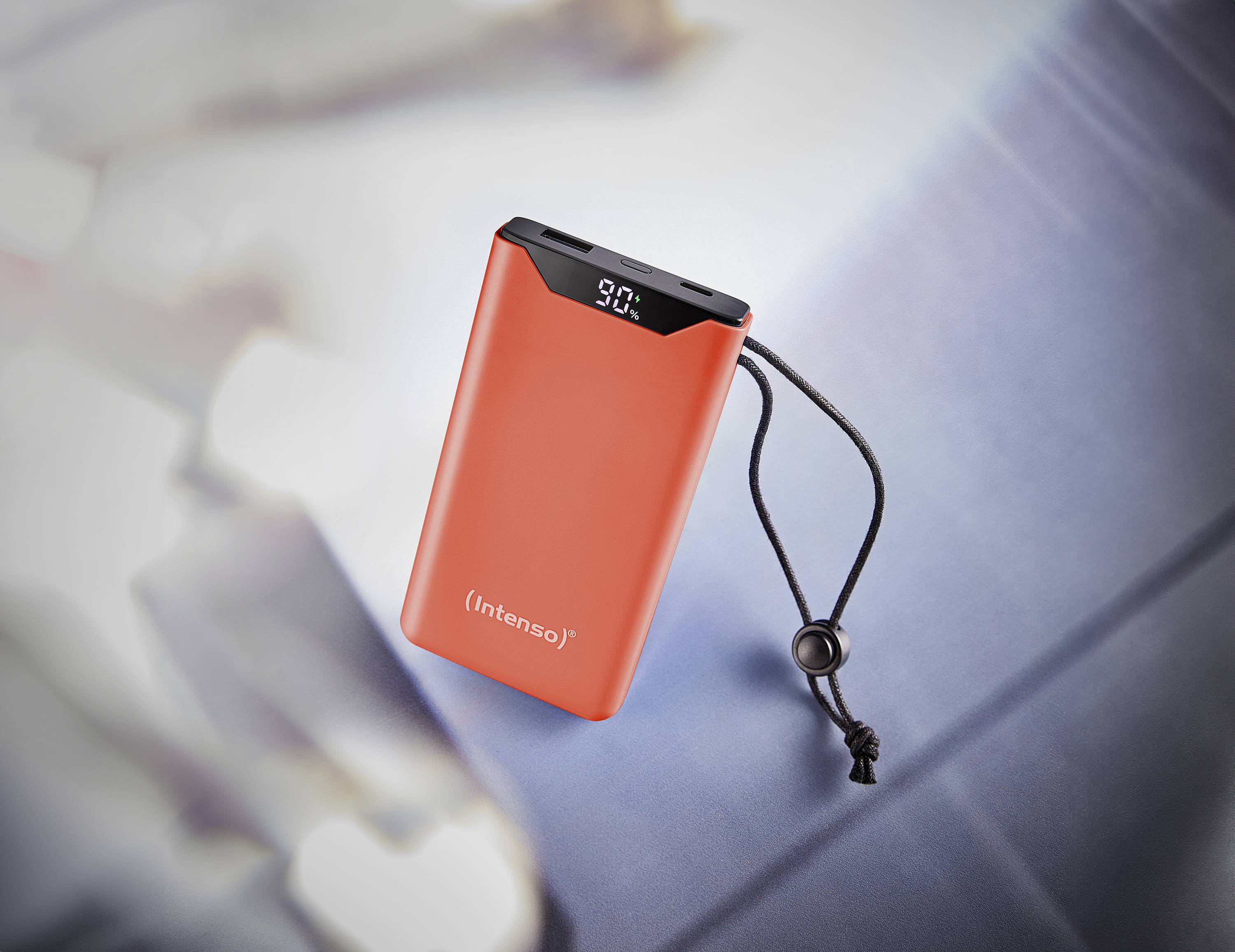 Rode powerbank met digitale weergave '90%' en zwarte draaglus, geplaatst op een metalen achtergrond.