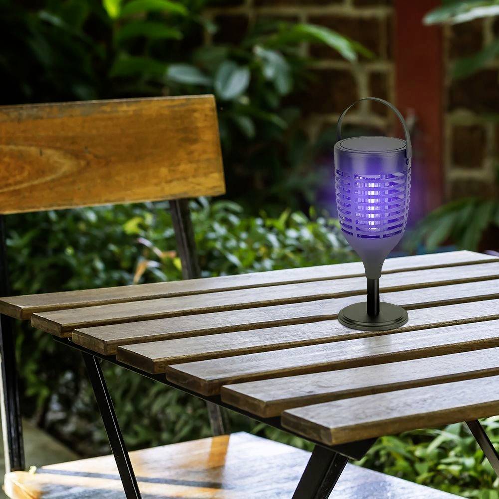 Een elektrische insectenverdelger staat op een houten tafel buiten, omringd door planten en een houten hek op de achtergrond.