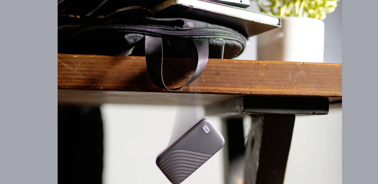 'Externe harde schijf valt van tafel' toont een draagbaar apparaat in vrije val naast een open laptoptas, plant op de achtergrond.