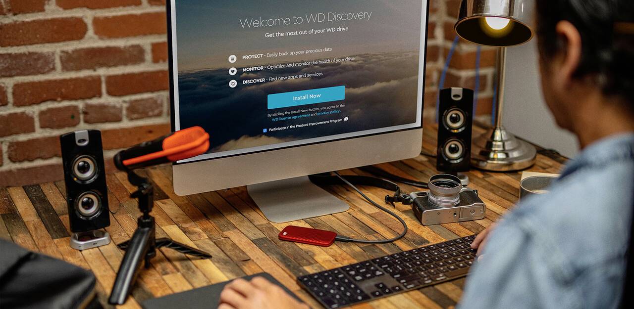 Een persoon werkt aan een bureau met een computer waarop het scherm 'Welcome to WD Discovery' weergeeft. Op het bureau staan speakers, een lamp, een camera en een externe harde schijf.