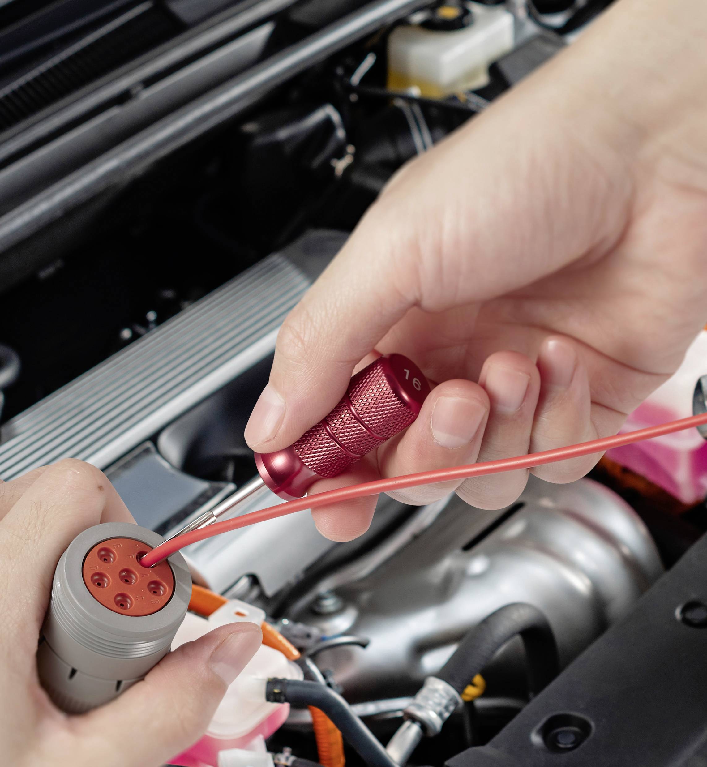 'Close-up van handen die een autobekabel verbinden met een rood gereedschap. Op de achtergrond is een motorruimte zichtbaar.'