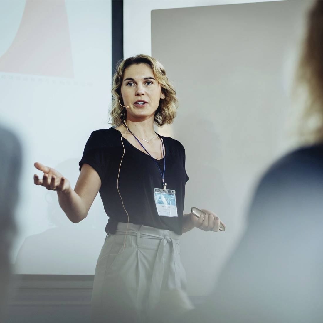Een vrouw geeft een presentatie in een vergaderzaal, draagt een headset en wijst naar een scherm met diagrammen op de achtergrond.