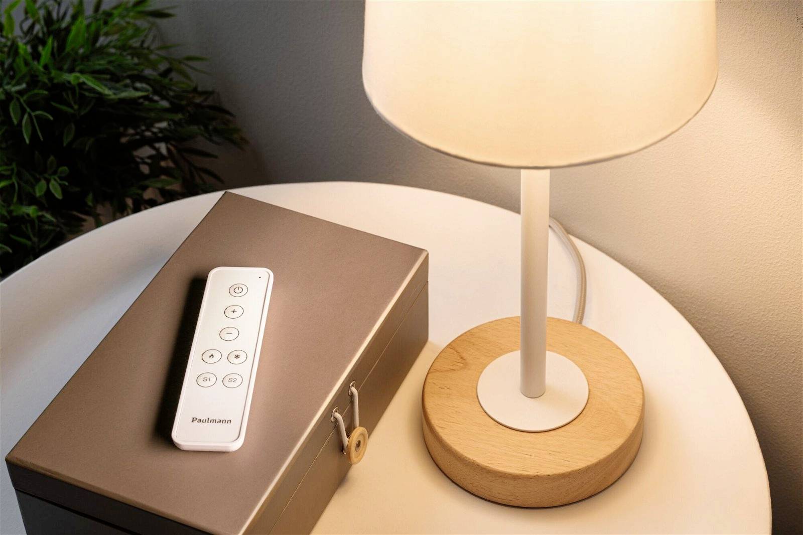 Witte afstandsbediening op een bruine doos naast een verlichte tafellamp op een rond tafeltje.