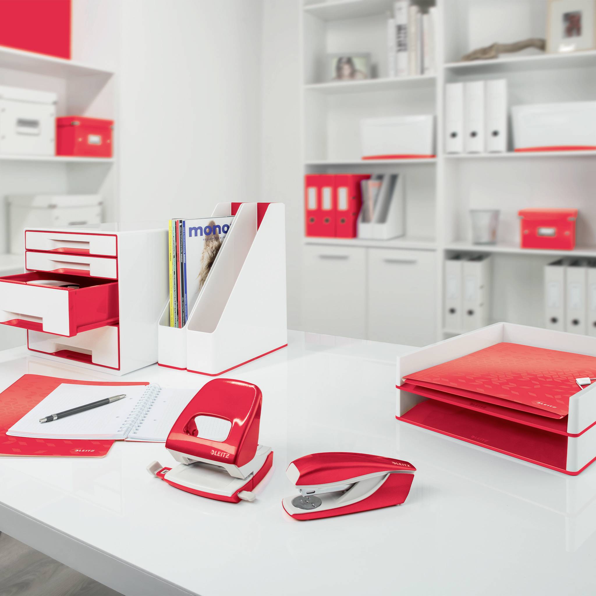 Een bureau met rood en wit kantoormateriaal, waaronder een nietmachine, perforator, map en notitieboek. Op de achtergrond planken met mappen.