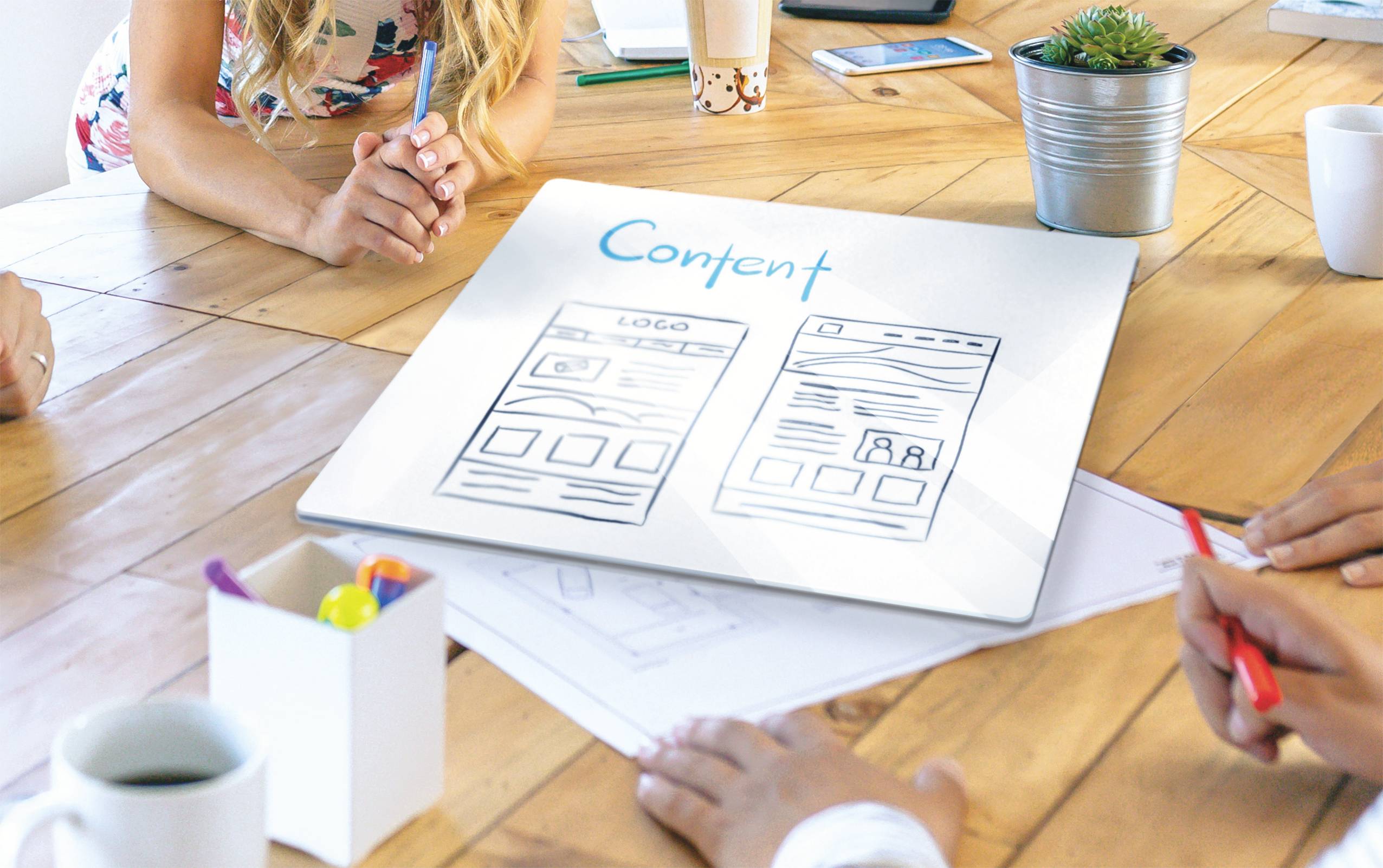 Mensen discussiëren over webdesign-schetsen op een tafel waarop het woord 'Content' staat. Gereedschappen en planten zijn zichtbaar.