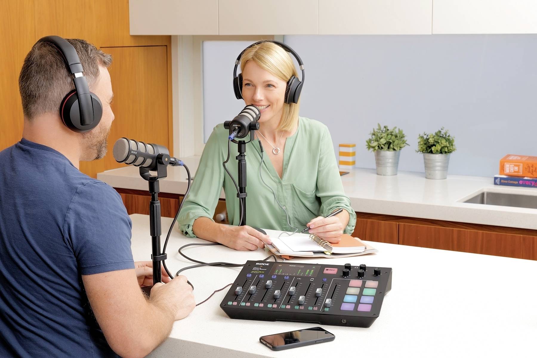 Twee personen met koptelefoons voeren een gesprek in een keuken, gebruiken microfoons en een audiomixer; een telefoon ligt ernaast.