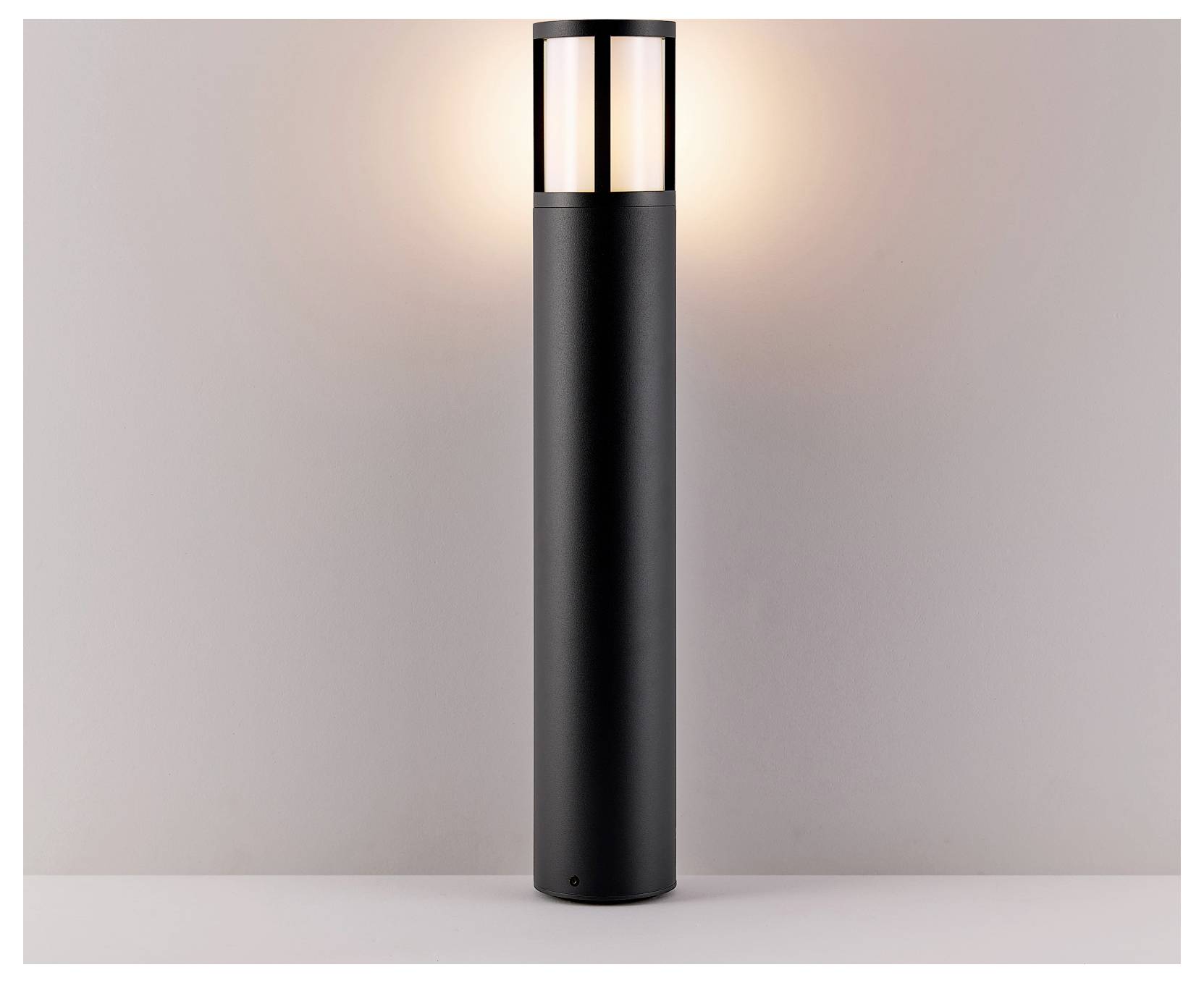 Zwarte cilindrische buitenlamp op een paal, die zacht oplicht tegen een effen muur.