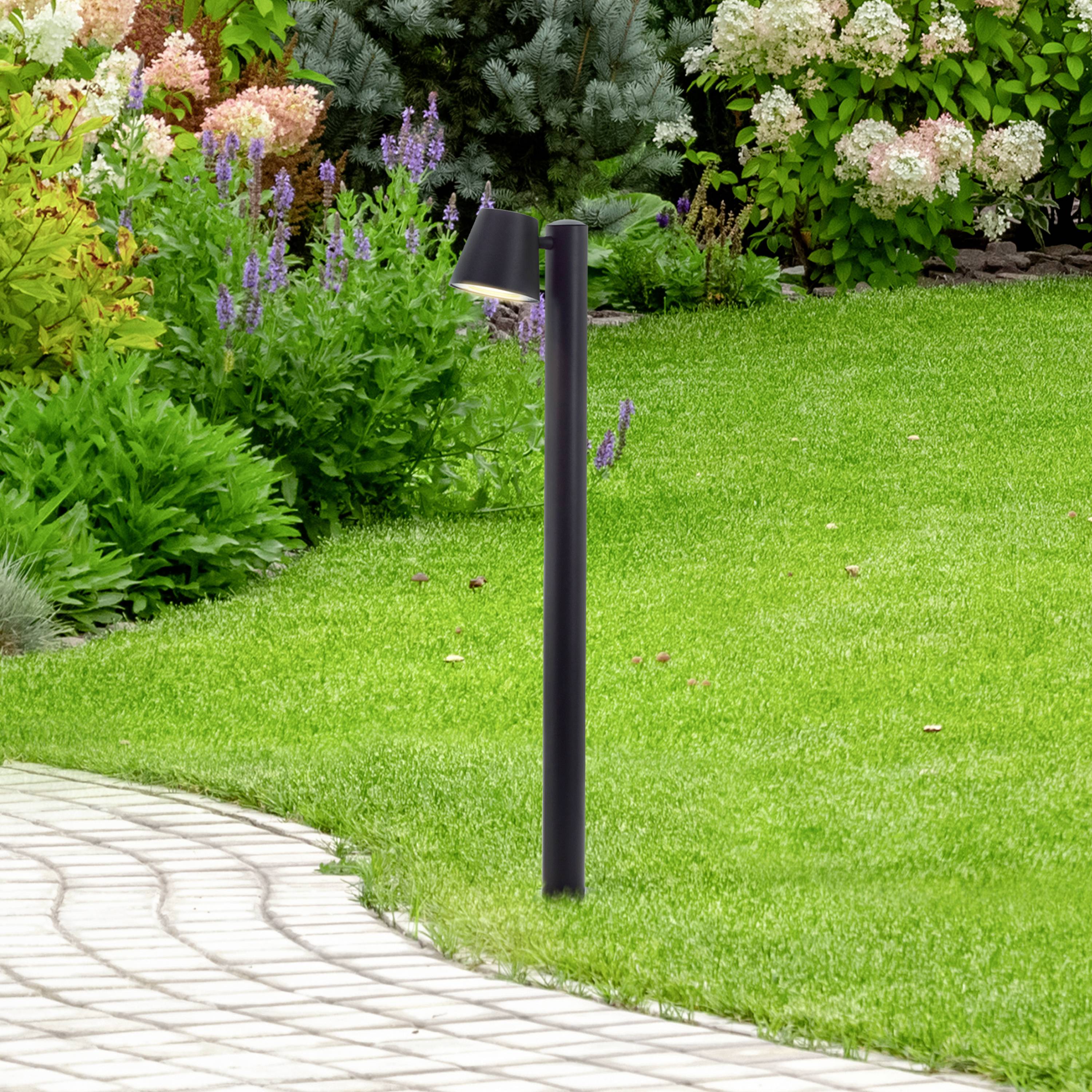 Tuin met groen gazon, geflankeerd door bloeiende planten en een slingerend bestratingspad. Een zwarte lantaarn staat op de voorgrond.
