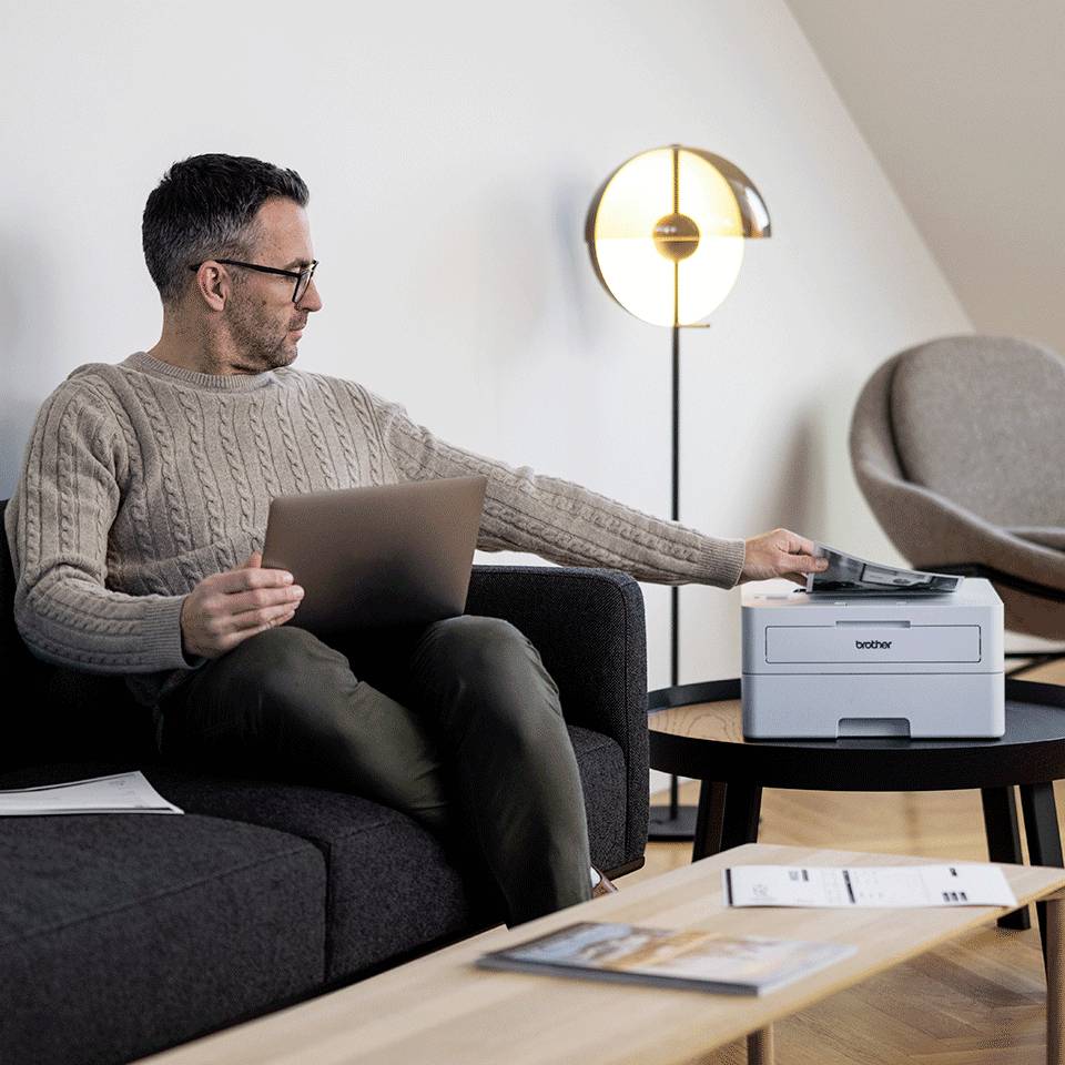 Een man zit op een bank en werkt op een laptop. Naast hem staat een printer op een tafel. Op de achtergrond is een staande lamp.