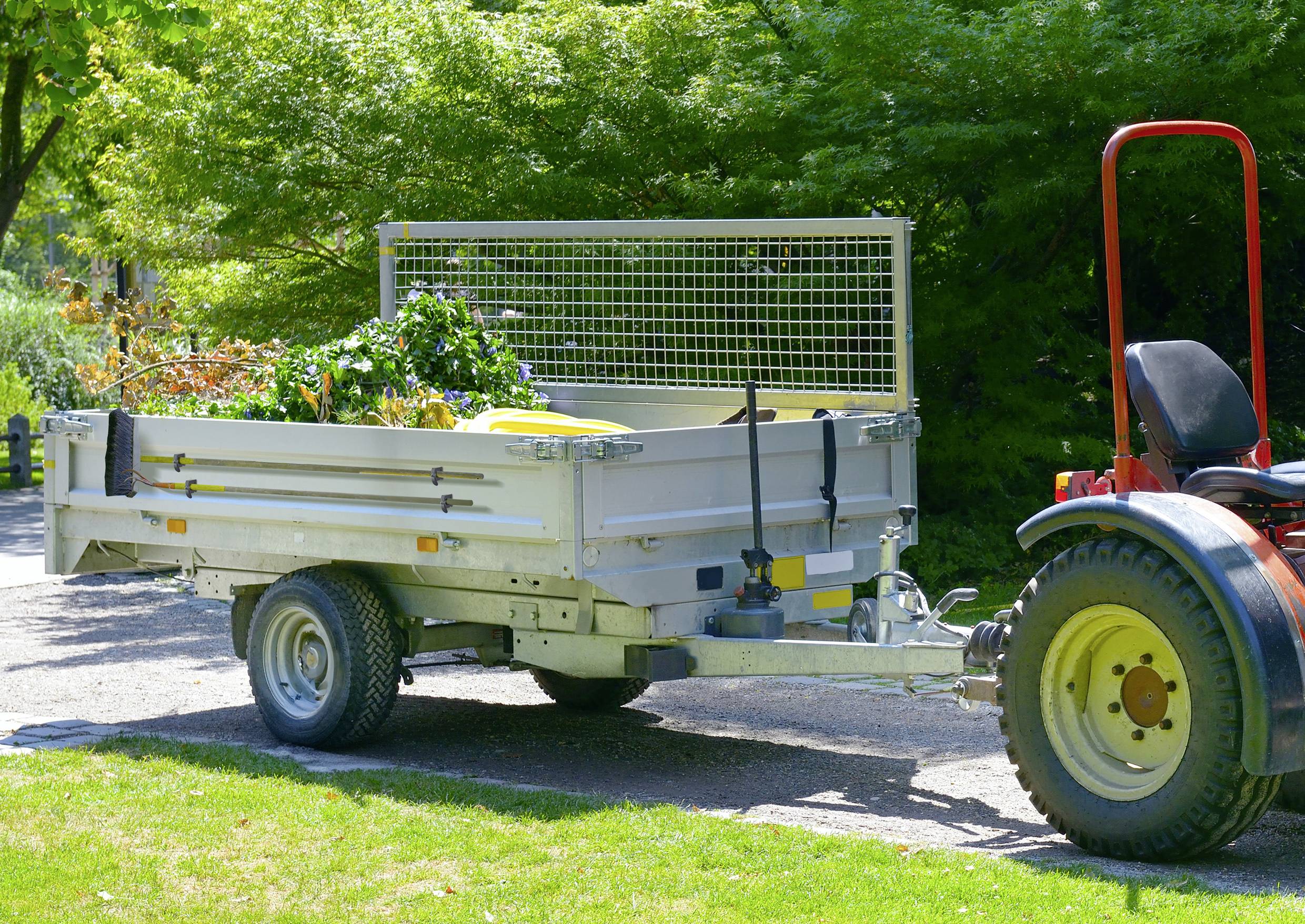 Een kleine tractor trekt een aanhanger vol tuinafval op een zonnig pad, omgeven door bomen op de achtergrond.