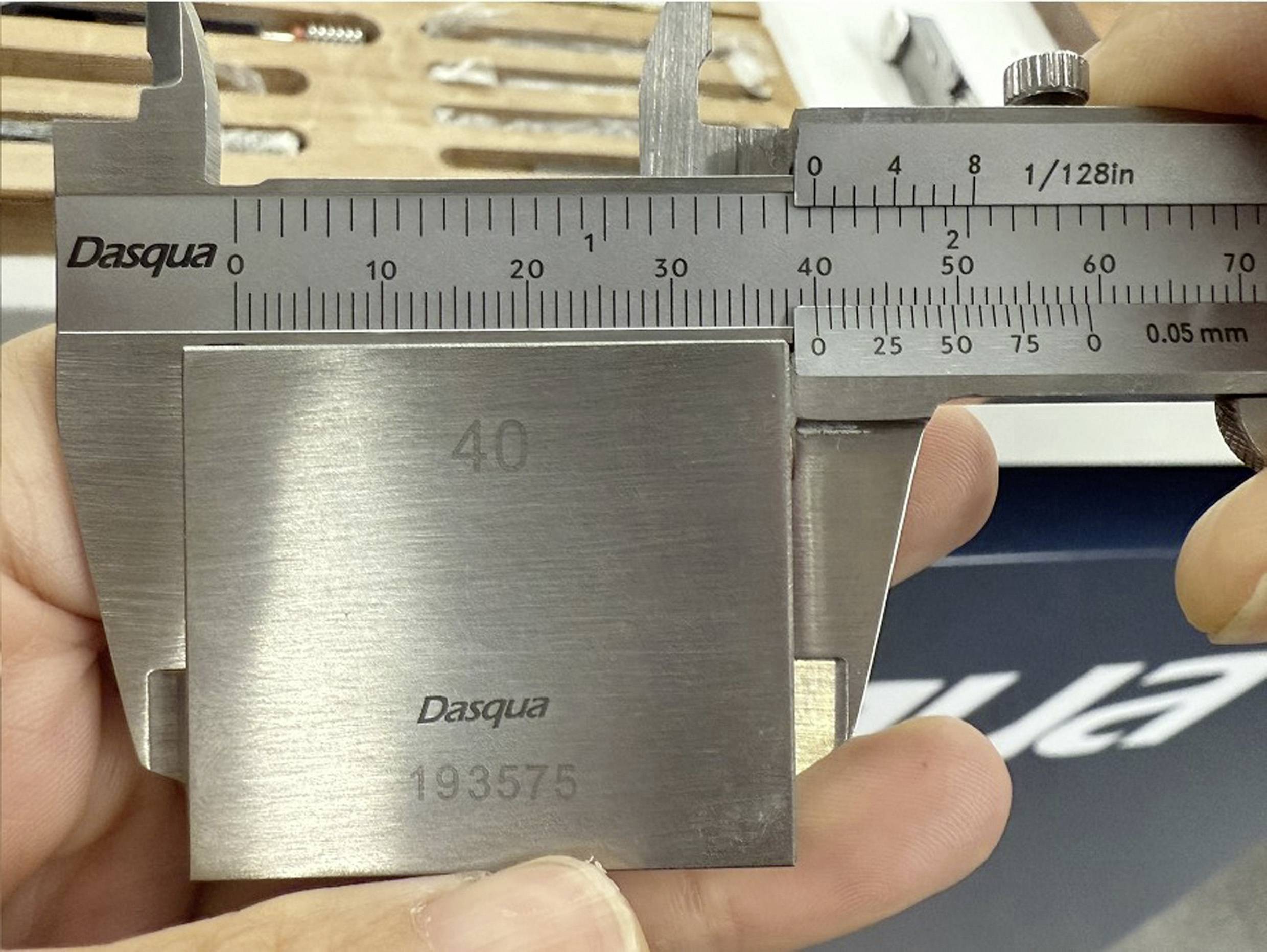 Een hand houdt een schuifmaat vast die de dikte van een metalen blok met opschrift '40' meet. De schuifmaat geeft ongeveer 40 mm aan.