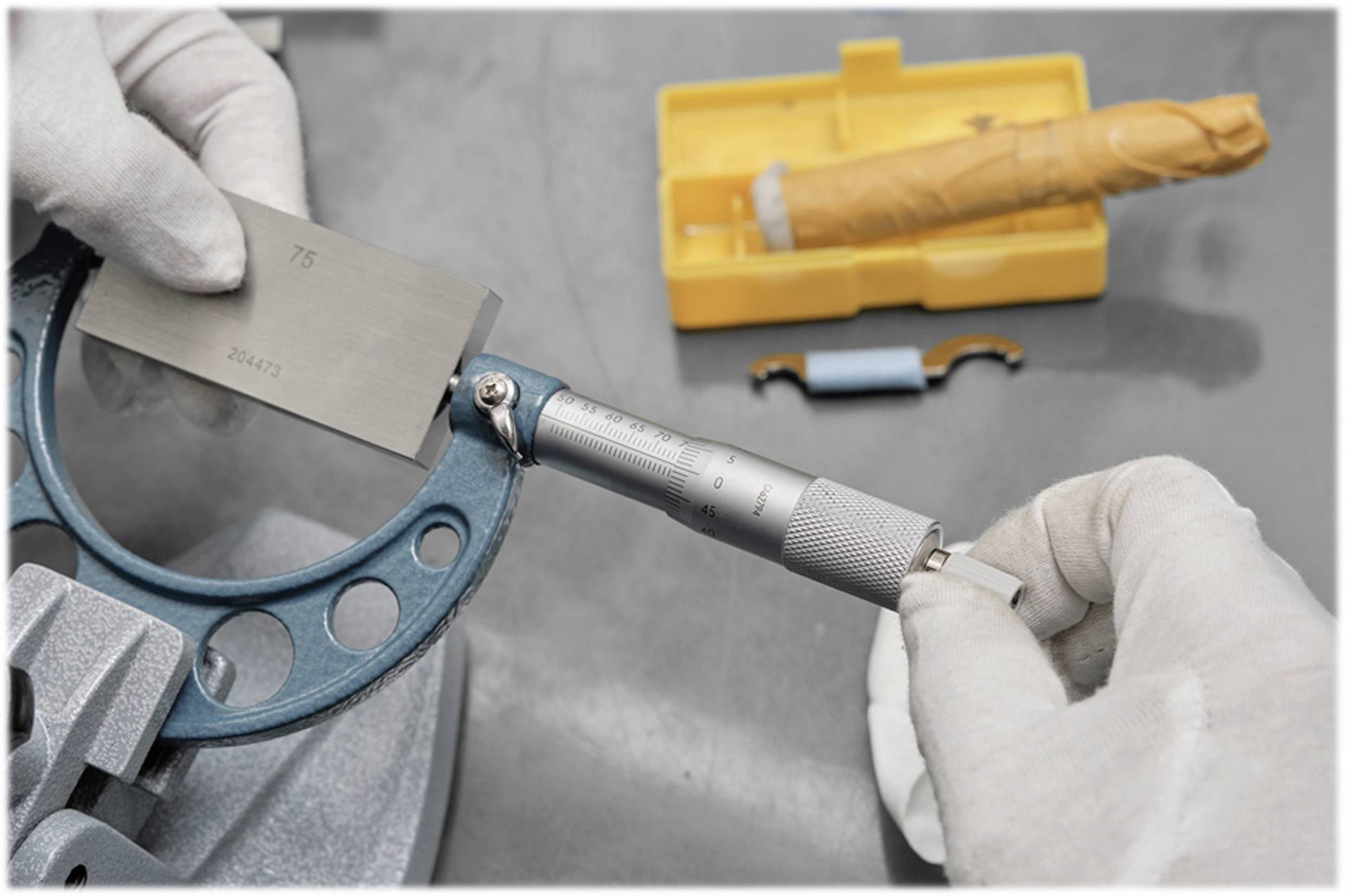 'Een persoon met handschoenen houdt een schuifmaat vast en meet een metalen onderdeel; op de achtergrond een geel etui met instrumenten.'