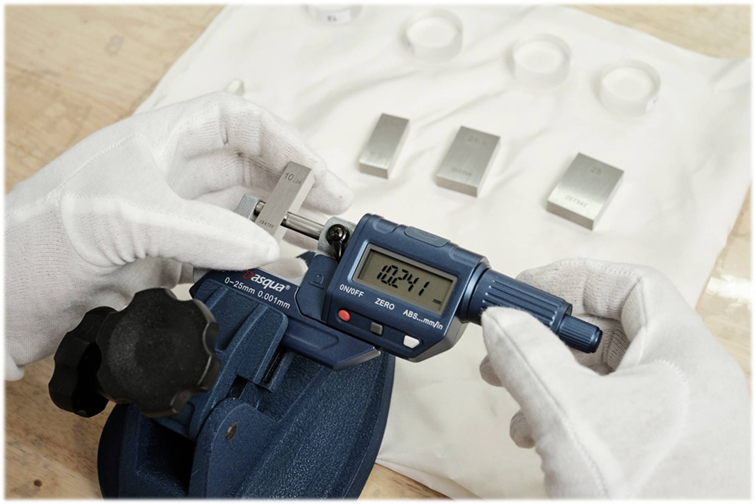 Een persoon meet een object met een digitale schuifmaat. Het display toont '102.41'. Op de achtergrond liggen metalen blokken.