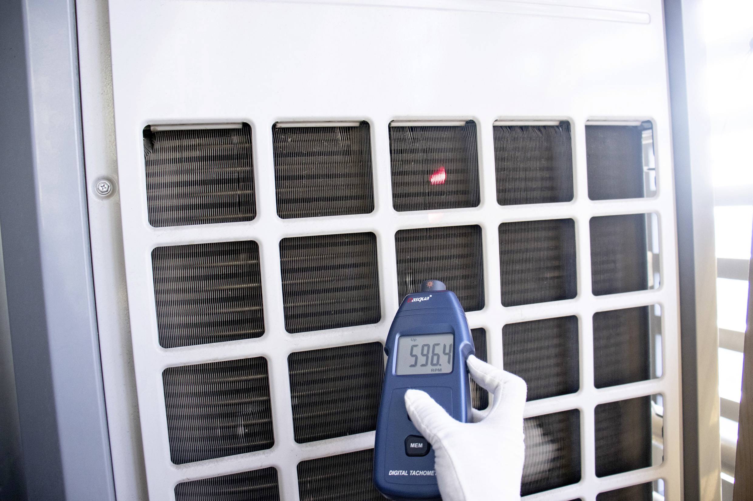 Een persoon meet de temperatuur van een airconditioning met een digitaal infrarood-thermometer. Het display toont '59,6 °C'.