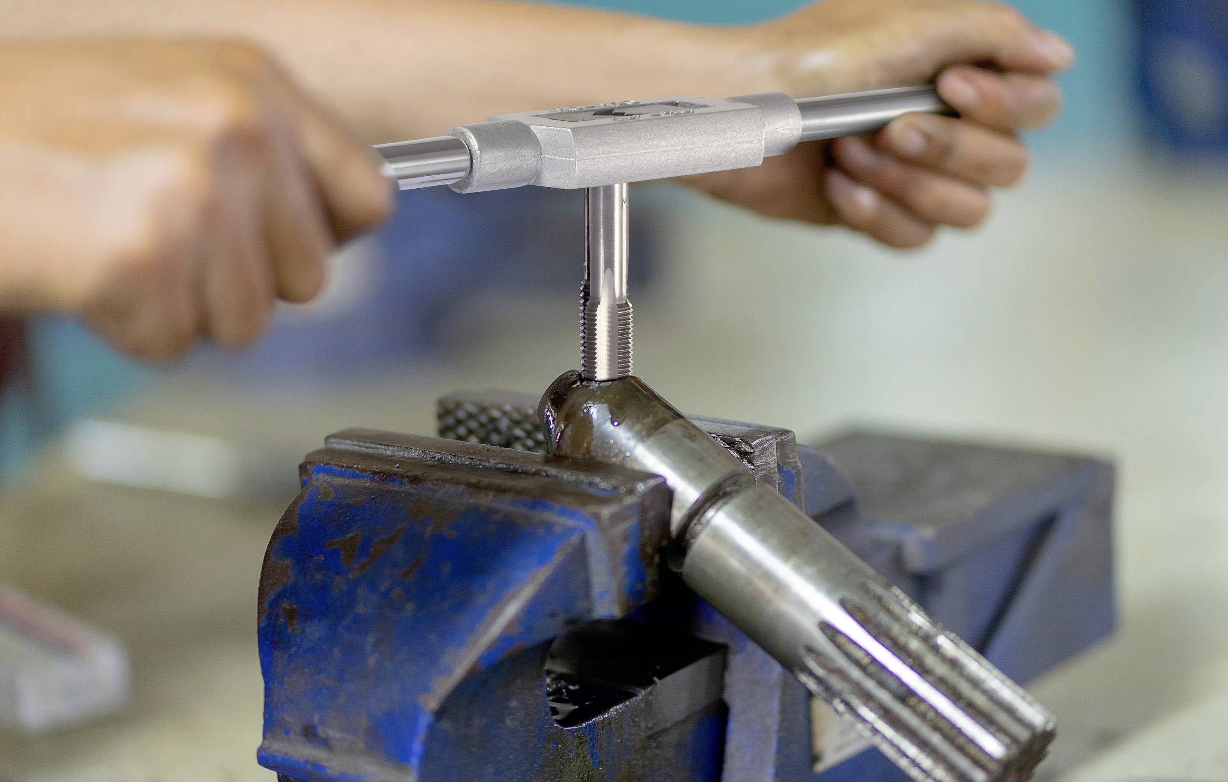 Een persoon gebruikt een draadtapgereedschap om schroefdraad te snijden in een metalen stuk dat vastgeklemd is in een bankschroef.