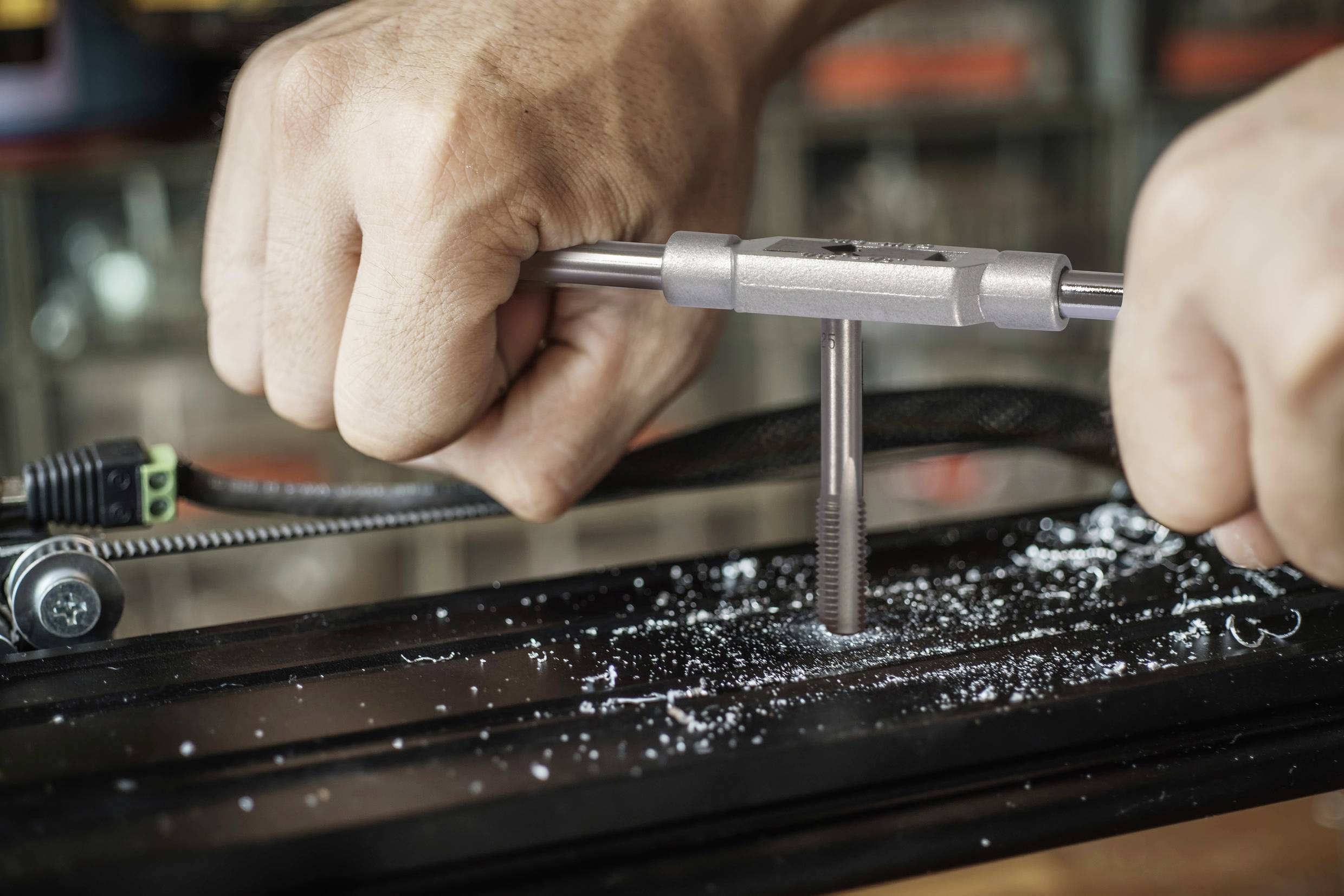 Twee handen snijden met een draadtap een nieuw schroefdraad in een metalen stuk. Metaalspanen zijn zichtbaar.