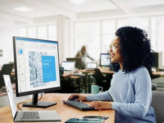 Een persoon werkt op een computer in een kantoor. Op het scherm is een presentatie te zien met een afbeelding van een gebouw.