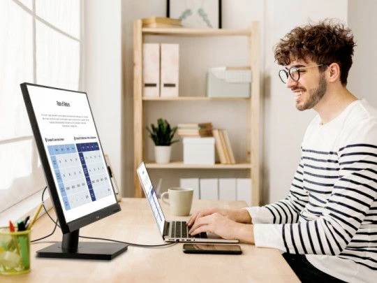 Jonge man met een bril werkt blij en opgewekt aan laptop en monitor in een licht kantoor, met op de achtergrond een boekenplank en ramen met gordijnen.