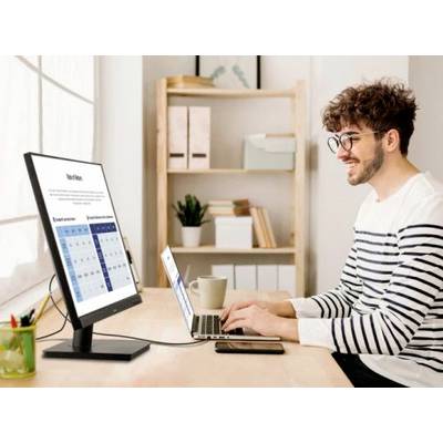 Jonge man met een bril werkt blij en opgewekt aan laptop en monitor in een licht kantoor, met op de achtergrond een boekenplank en ramen met gordijnen.