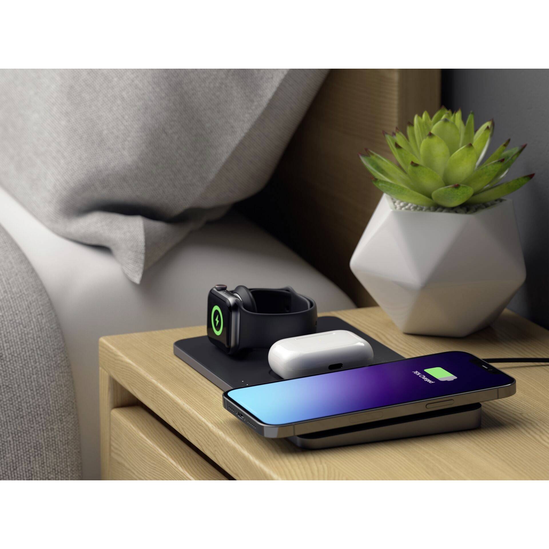 Een nachtkastscène met een laadstation dat een smartphone, smartwatch en koptelefoon oplaadt. Naast het laadstation staat een plant.