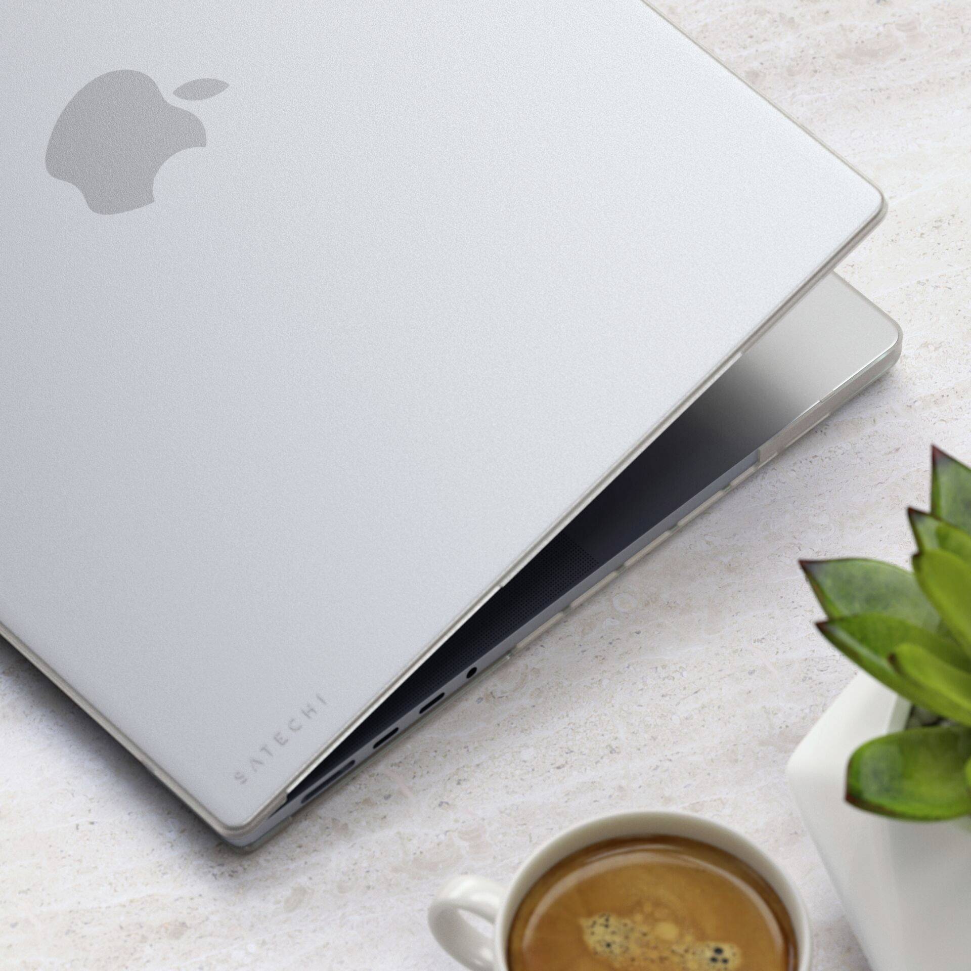 Een zilveren laptop met Apple-logo half geopend op een tafel naast een kopje koffie en een kleine plant.