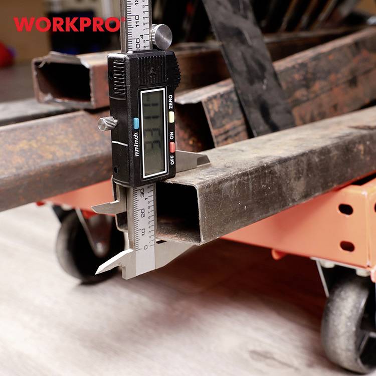 Digitale schuifmaat meet rechthoekig metalen buis op een werkbank. Op de achtergrond zijn houten pallets zichtbaar.