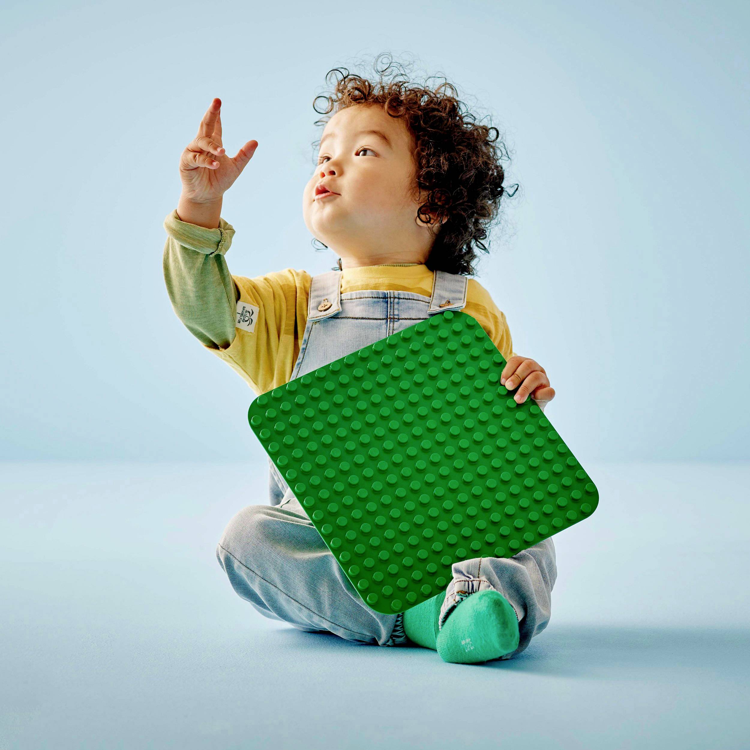 Kind zit op de grond, kijkt omhoog en houdt een grote groene bouwplaat vast. Draagt een geel shirt en een overall. Pastelblauwe achtergrond.