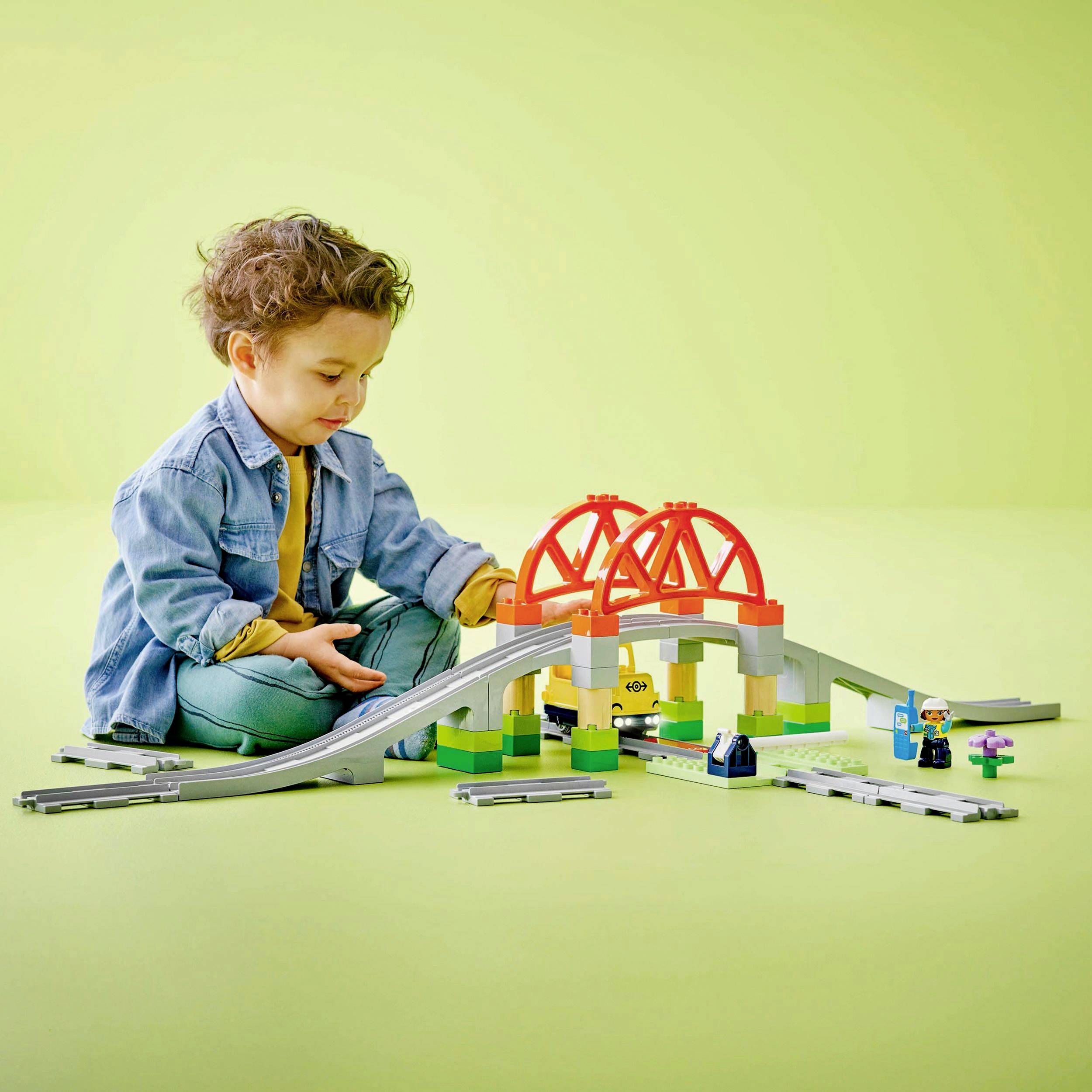 Een kind speelt met een kleurrijke speelgoed-spoorbrug op een groene ondergrond. Het draagt een spijkerjas en een geel shirt.