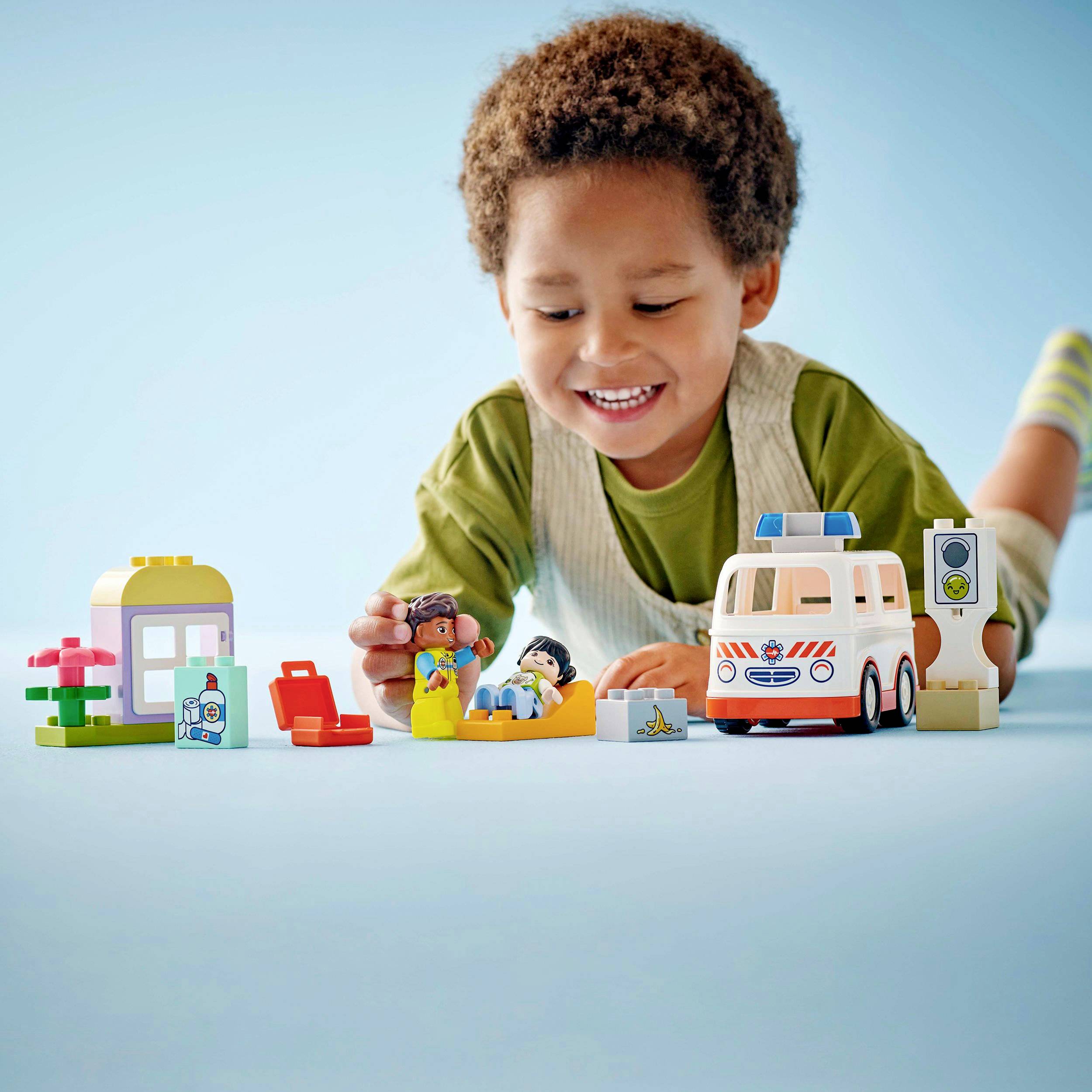 Een lachend kind speelt met LEGO-blokken, waaronder een ambulance en een arts. Op de achtergrond is een LEGO-huis te zien.